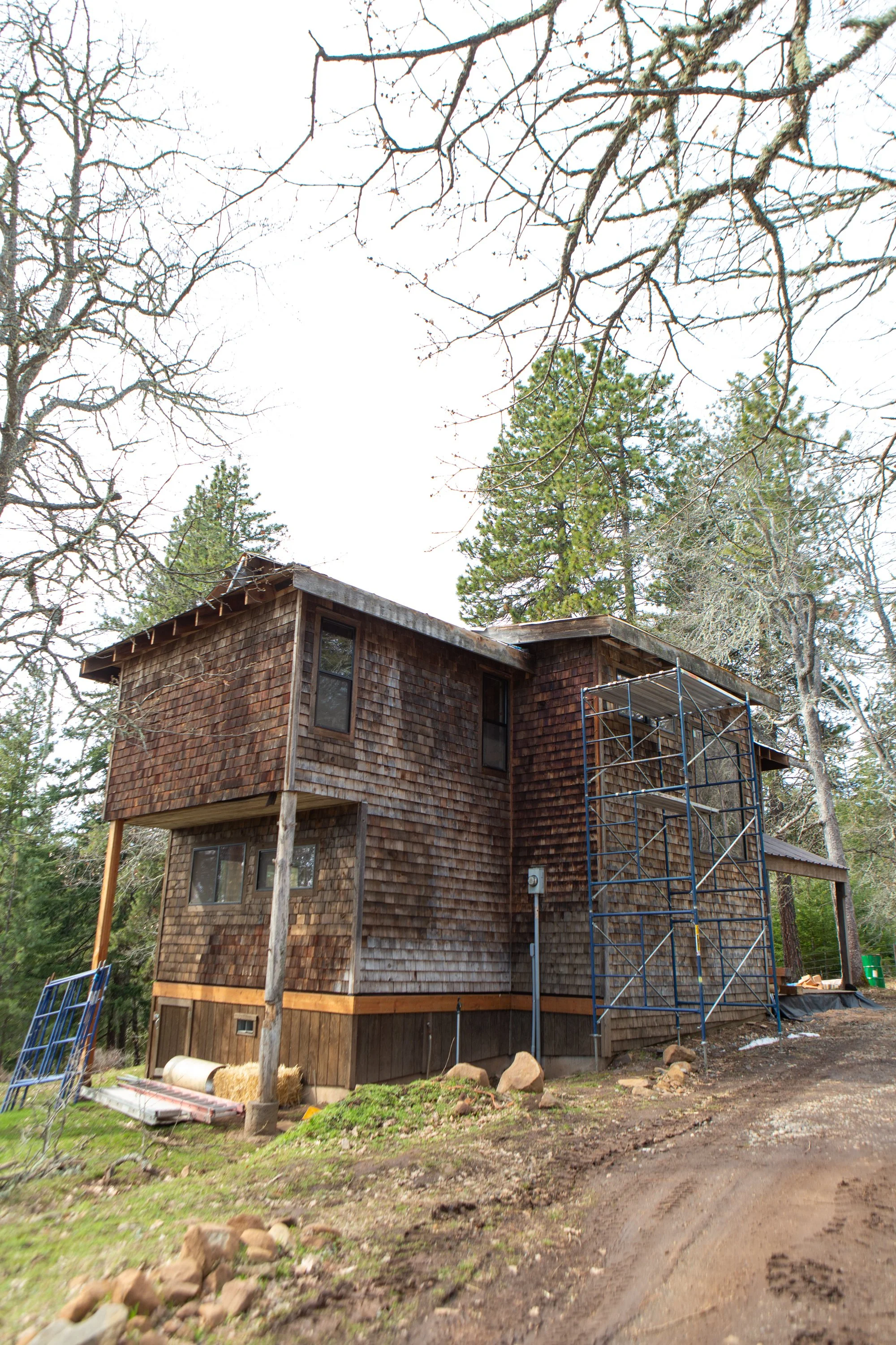 A two-story wooden house under construction with scaffolding on the side, surrounded by trees and a dirt road.