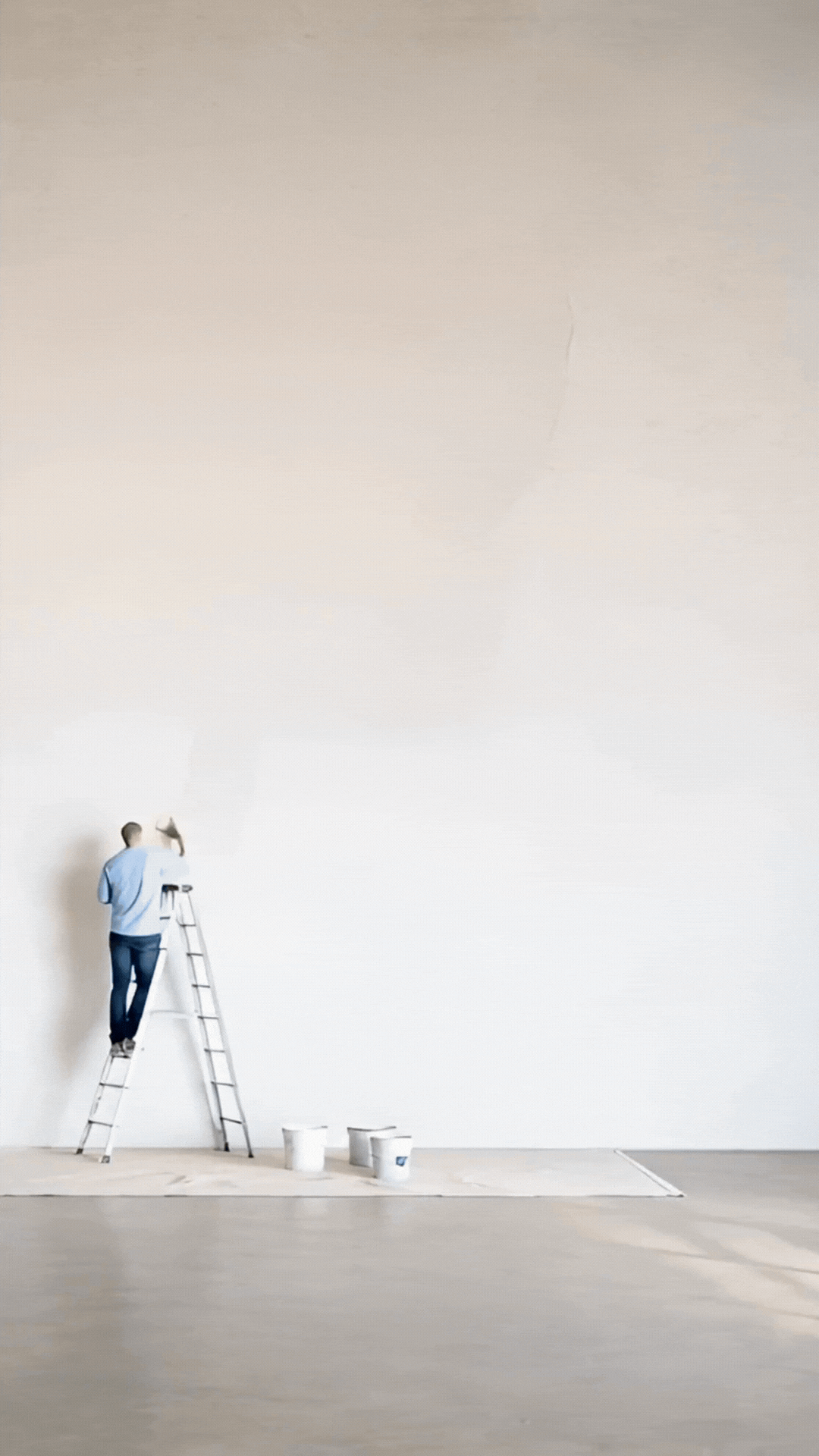 A person standing on a ladder painting a large, blank white wall in a room with a paint bucket and a drop cloth on the floor.