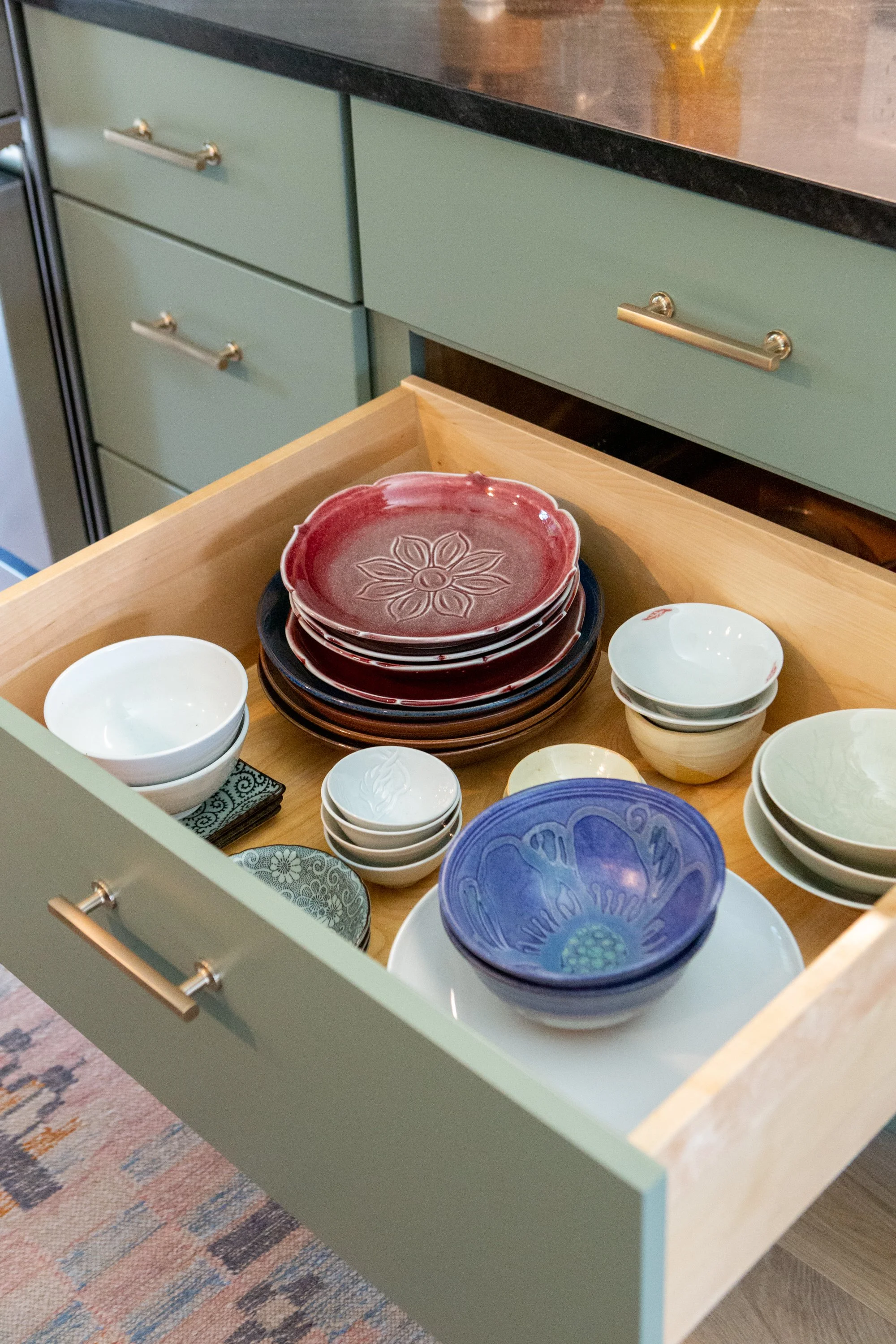 Open drawer containing colorful ceramic bowls with floral and abstract patterns, some stacked, in a kitchen with green cabinets and a black countertop.