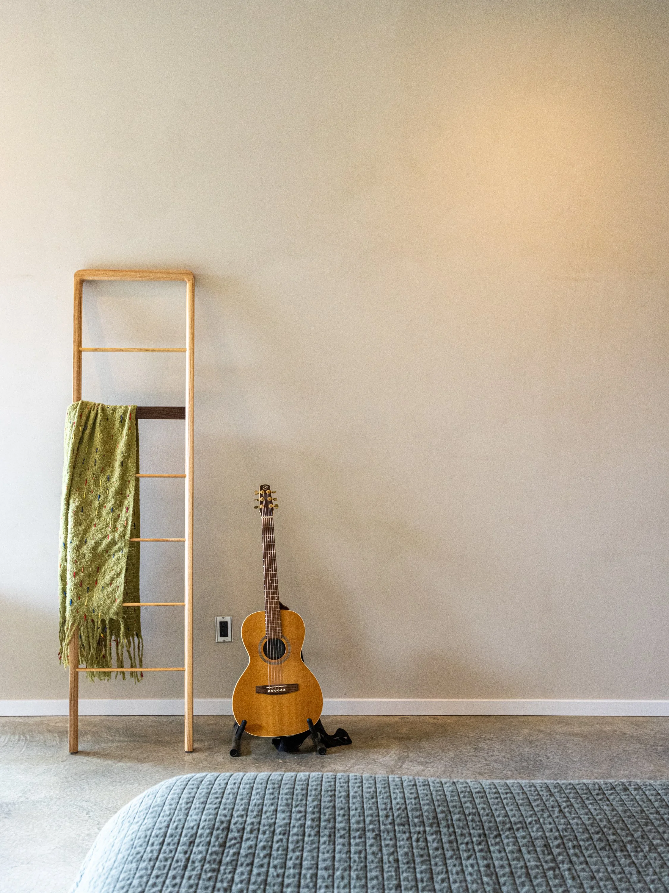 A room with a beige wall and carpeted floor. On the left is a wooden ladder with a green textured throw blanket hanging from one of the rungs. To the right is an acoustic guitar standing on a guitar stand. There is a wall outlet near the guitar.