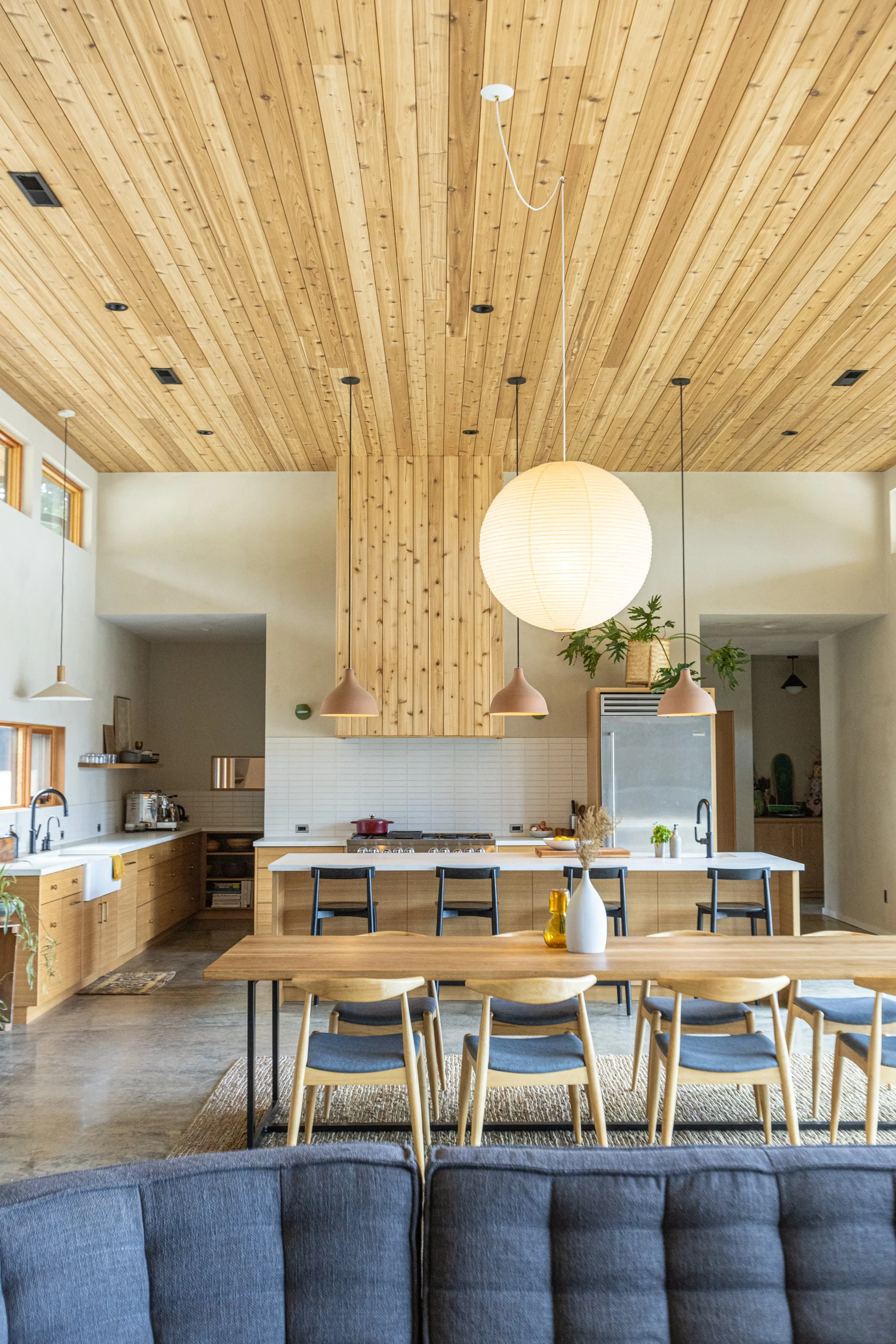 Modern kitchen with wooden ceiling and island, pendant and ceiling lights, dining table with chairs, and decorative vases.