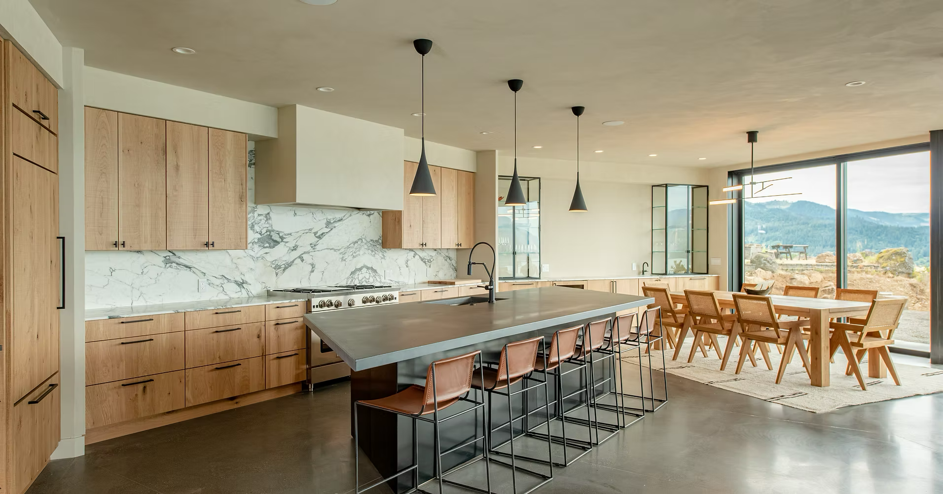 Open kitchen and dining area with natural wooden cabinets, marble backsplash, large island with black sink, modern pendant lights, and a dining table with eight chairs. Large glass sliding doors reveal an outdoor mountain landscape.
