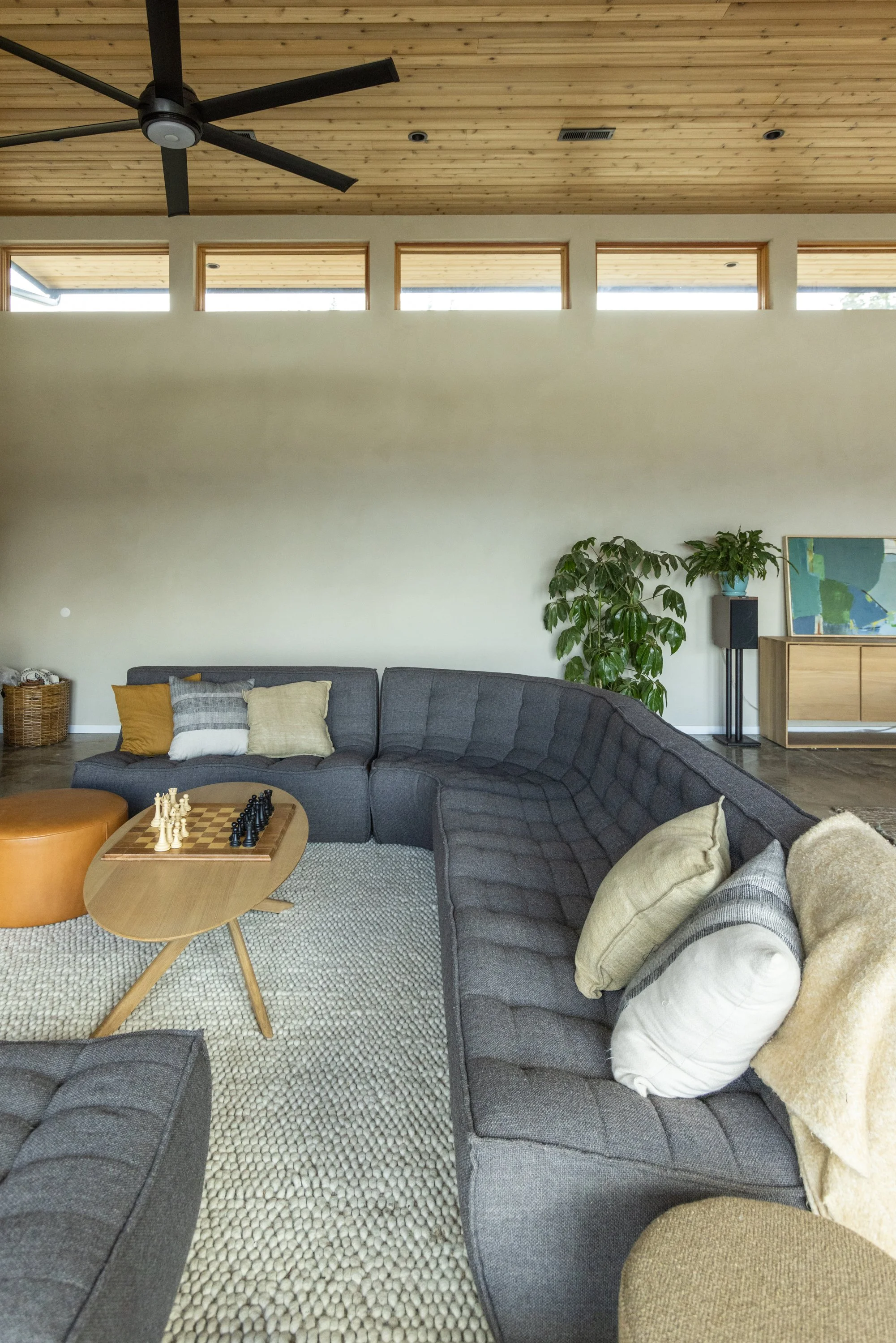 Living room with a dark gray sectional sofa, beige pillows, a wooden round coffee table with a chess board, potted plants, and a wooden sideboard with artwork and a speaker. The ceiling has a wooden finish with square windows and a black ceiling fan.