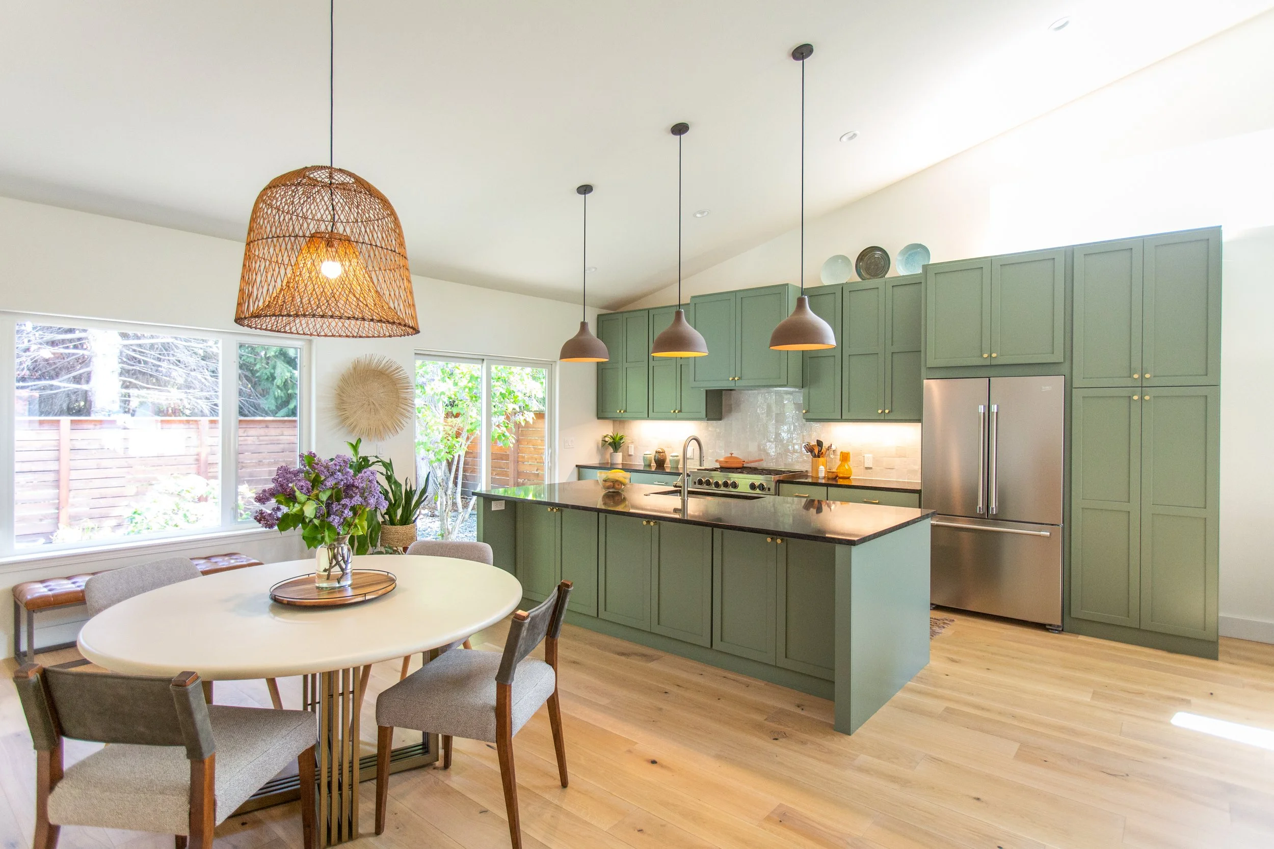 A bright kitchen with green cabinets, a large island, stainless steel appliances, pendant lighting, and a dining area with a round table, chairs, and a floral centerpiece.