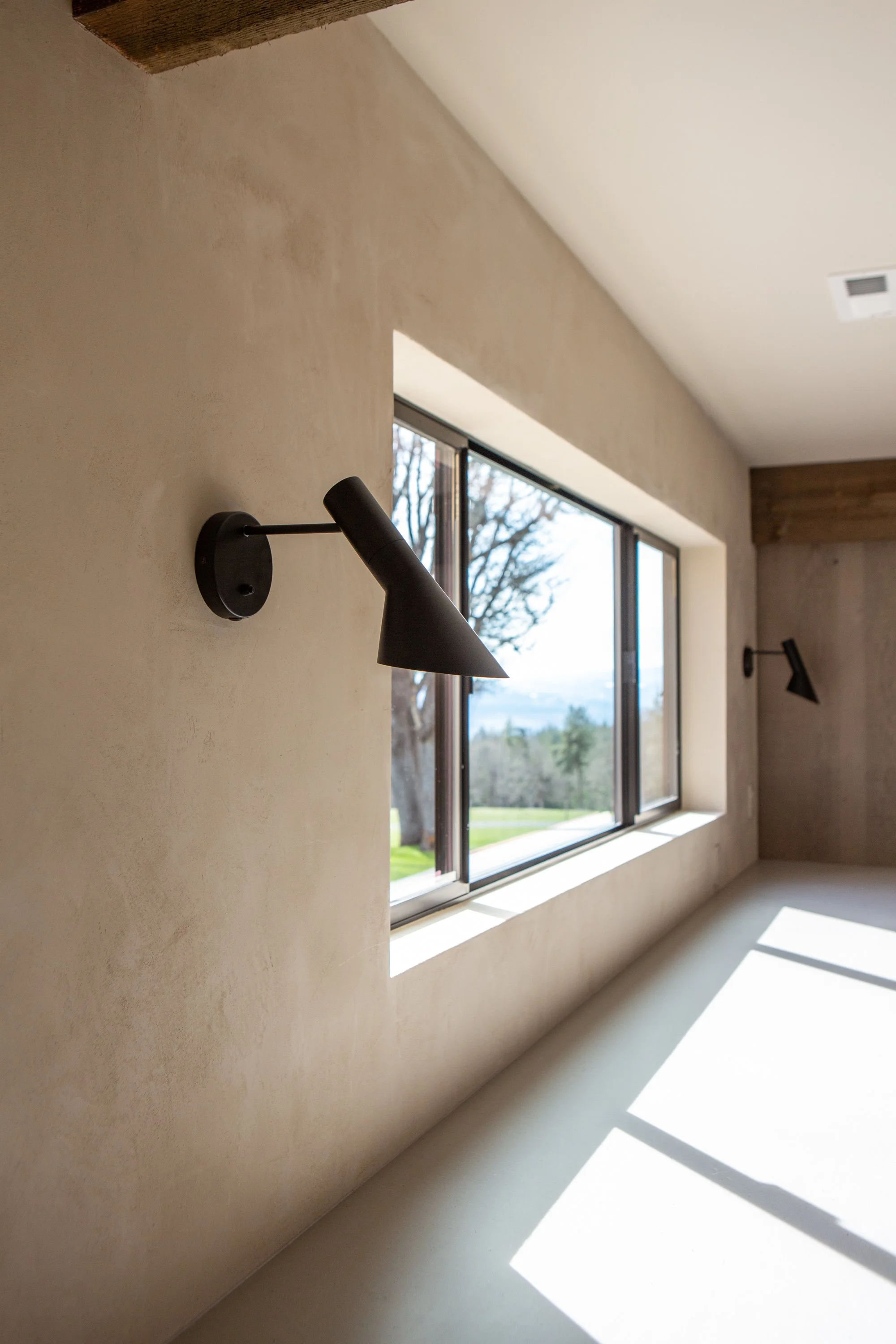 A minimalist interior room with a large window allowing natural light to cast shadows on the floor, a wall-mounted black reading lamp, and view of trees outside.