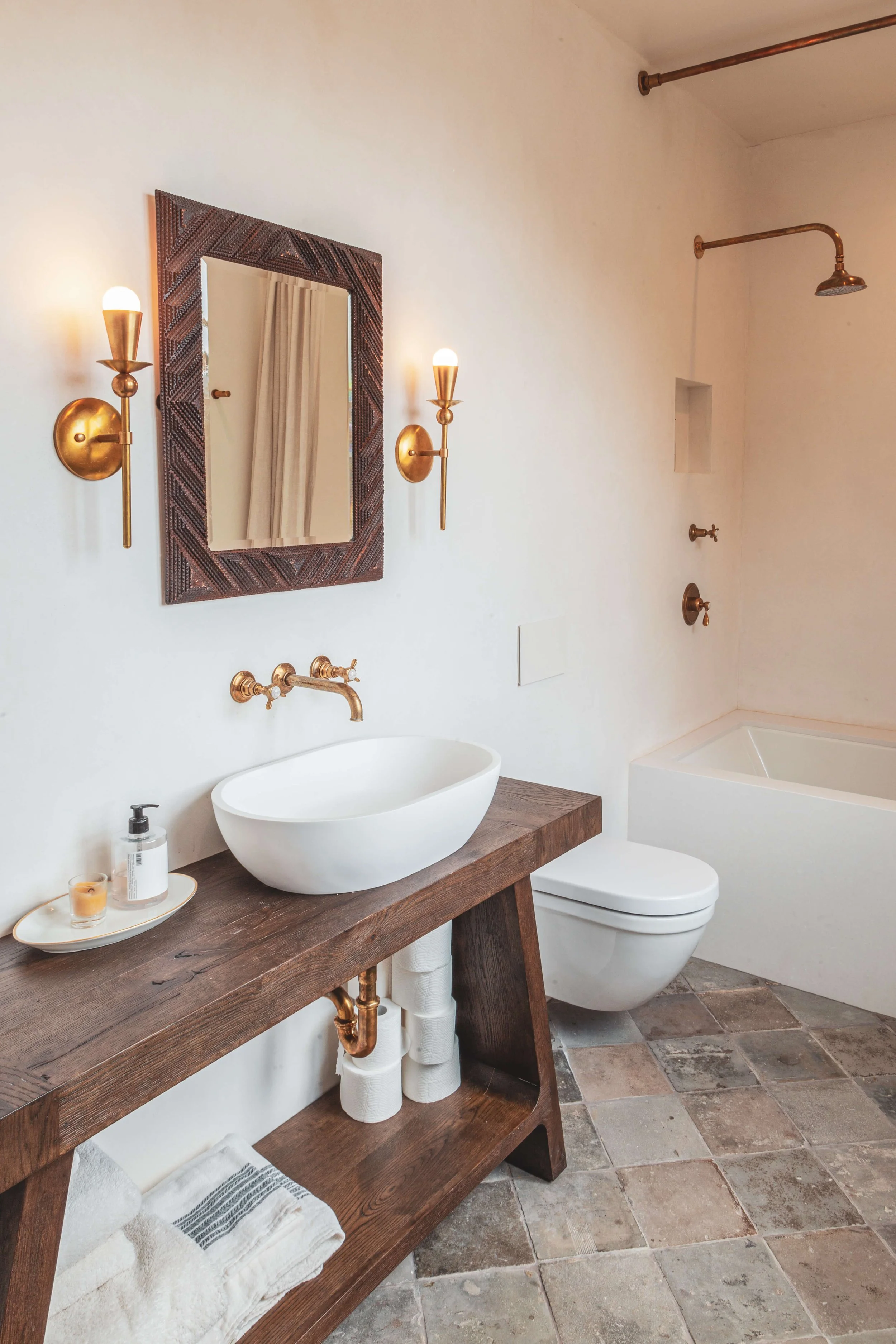 Modern bathroom with a wooden vanity, vessel sink, gold fixtures, gold wall sconces, a mirror with a decorative frame, a bathtub, a toilet, and a shower with a brass showerhead, on a tiled floor.