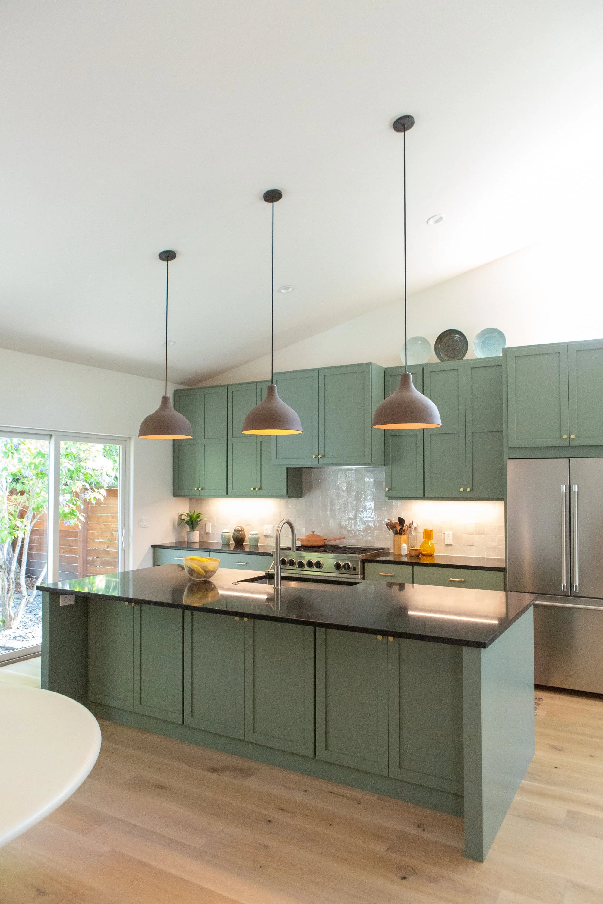 Modern kitchen with light green cabinets, black countertops, a kitchen island, hanging pendant lights, stainless steel appliances, and large windows letting in natural light.