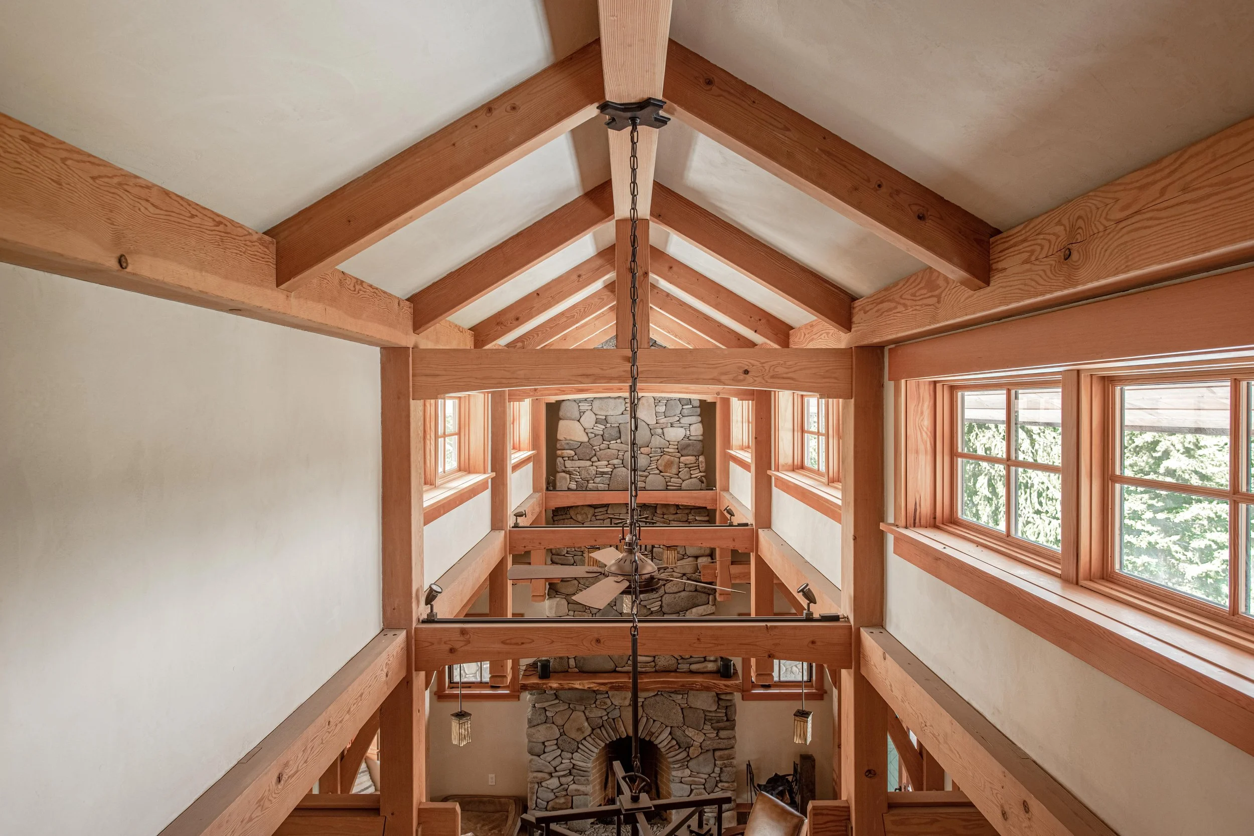 Interior view of a wooden house ceiling with exposed beams, windows, and a stone fireplace in the background.