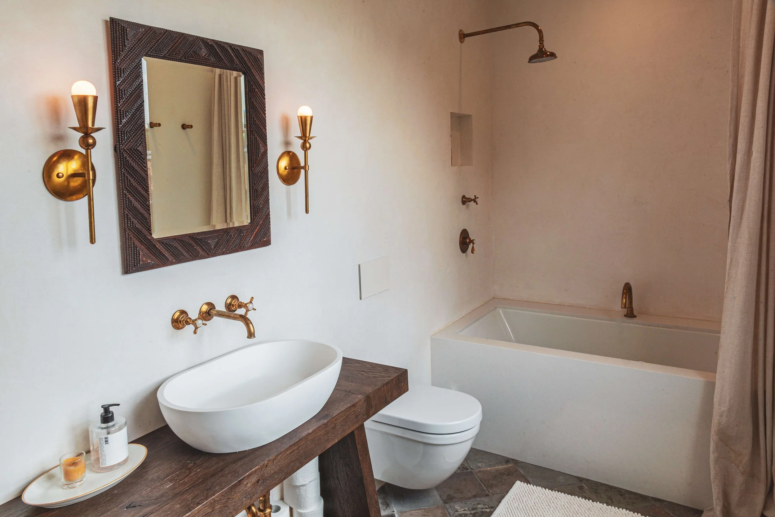 A modern bathroom with a white oval bathtub, a wooden vanity with a white vessel sink, gold wall-mounted light fixtures, a mirror with a textured frame, a shower area with a gold showerhead, a toilet, and a small shelf built into the wall. The floor 