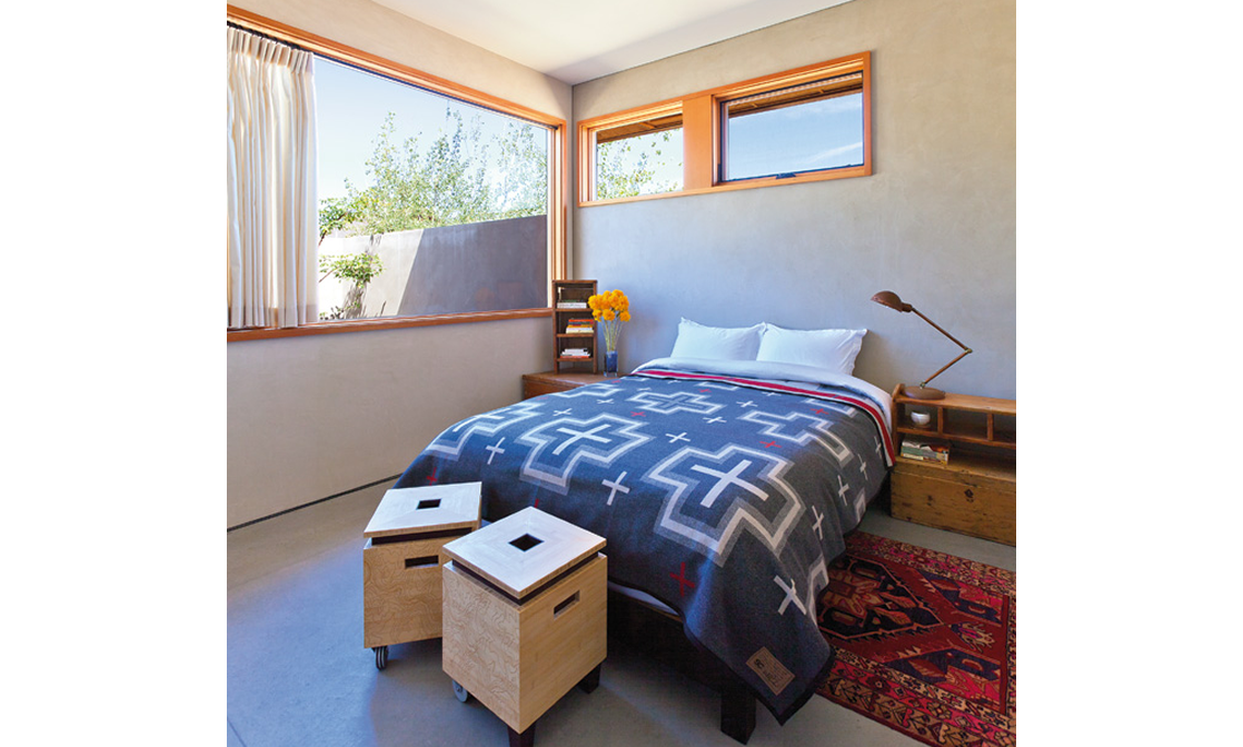 Modern bedroom with large corner window, bed with patterned blanket, small wooden bedside table, vintage-style lamp, and two wooden storage crates on wheels.