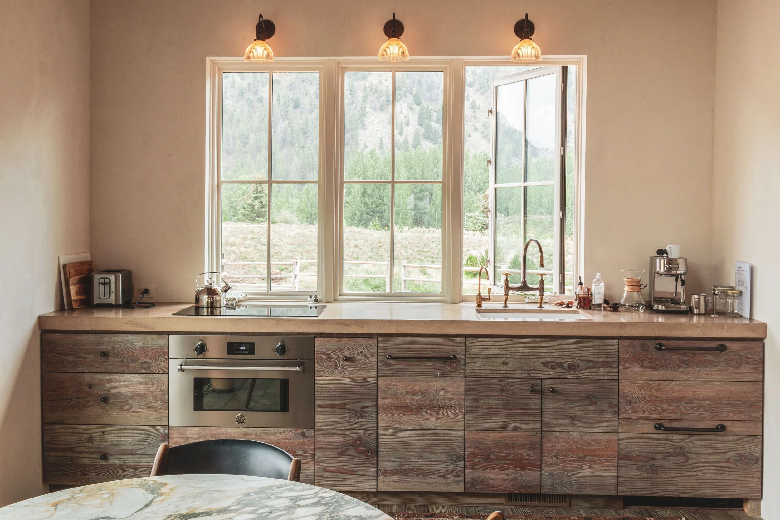 A kitchen with a large window above the countertop, showing a mountain landscape outside. The countertop has kitchen appliances like a toaster, kettle, and coffee maker, with a built-in oven below. There are three pendant lights hanging from the ceil