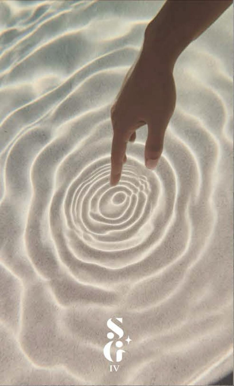 A person's hand touching the sand close to the shore creating ripples in the water.