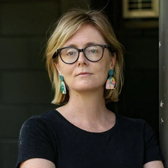A woman with shoulder-length blonde hair, wearing black glasses, colorful earrings, and a black shirt. She appears to be outdoors in front of a dark background.