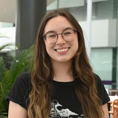 Smiling young woman with long brown hair, glasses, and a black T-shirt standing outdoors with modern buildings and greenery in the background.