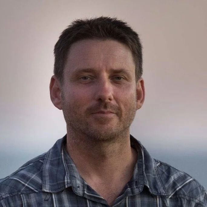 A man with short brown hair, slight beard, wearing a blue plaid shirt, standing outdoors with a blurred sky background.