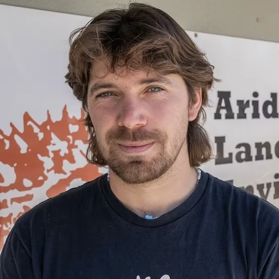 A young man with light brown, slightly wavy hair and a beard, wearing a dark t-shirt, standing outdoors in front of a sign with partial text and an orange design.