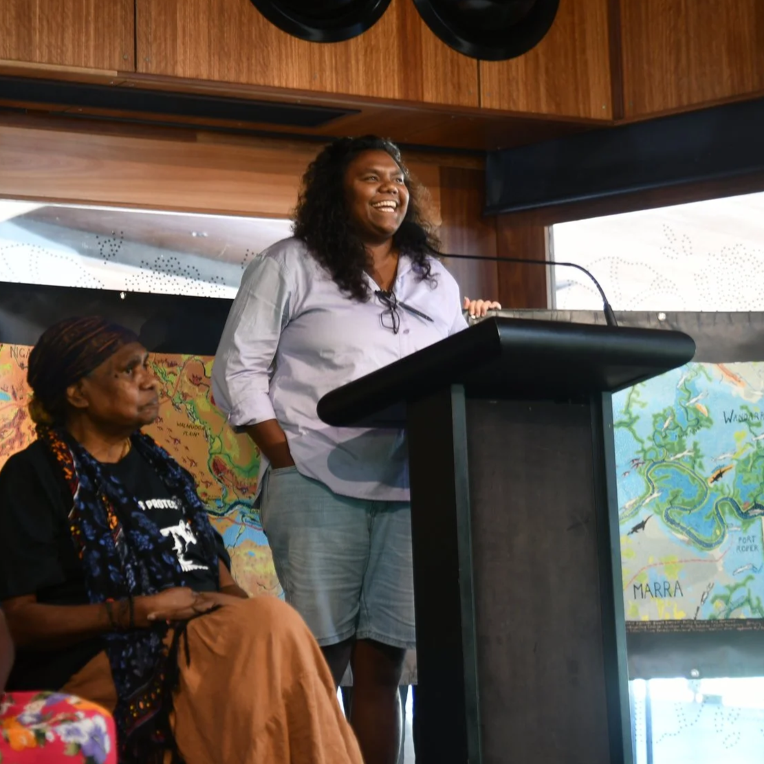 A woman standing and speaking at a podium in a room with wooden walls and large windows, with two large maps on the wall behind her. Another woman is seated beside her, listening.