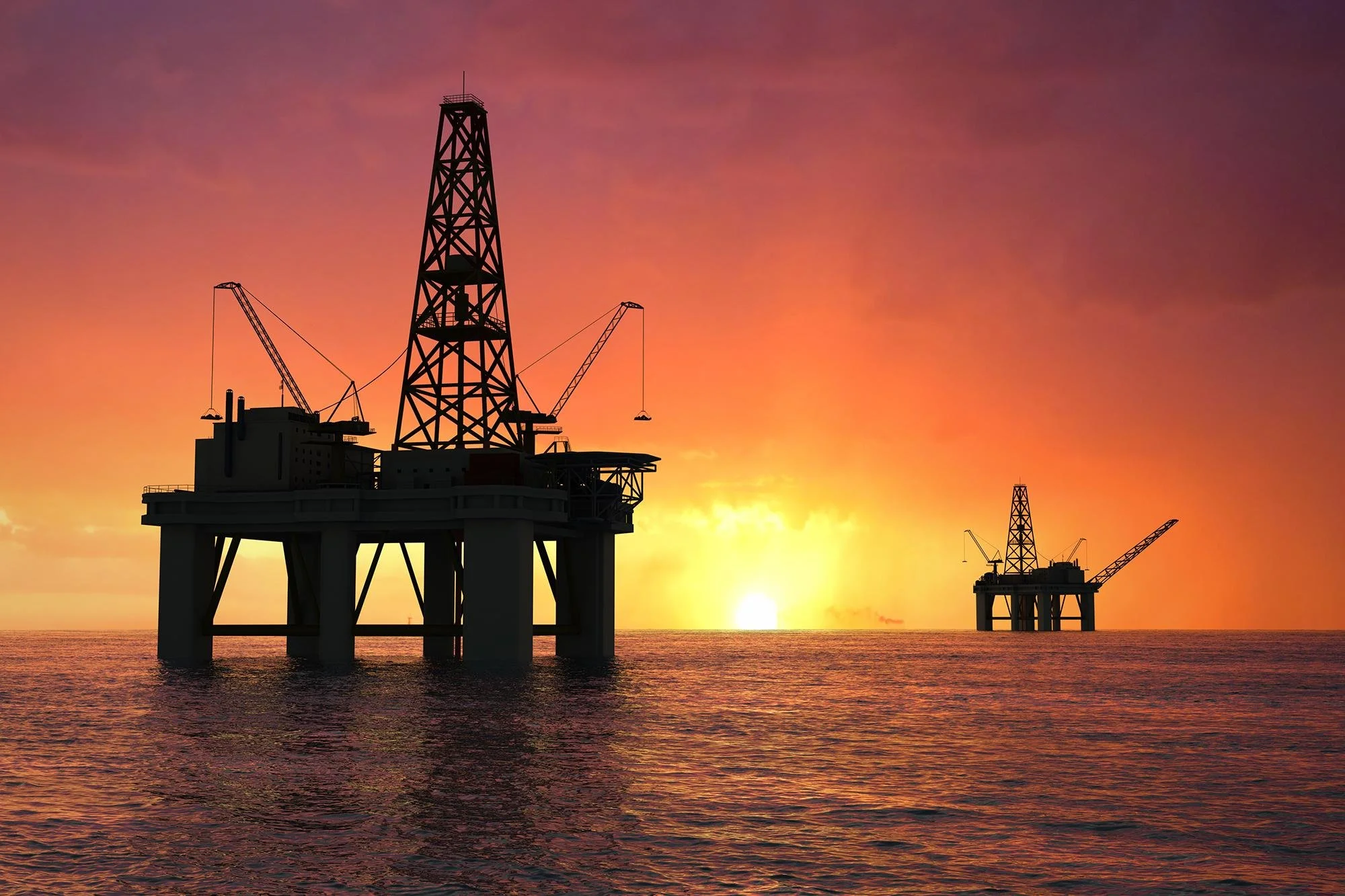Silhouettes of offshore oil rigs in the ocean at sunset with a colorful sky and calm water below.