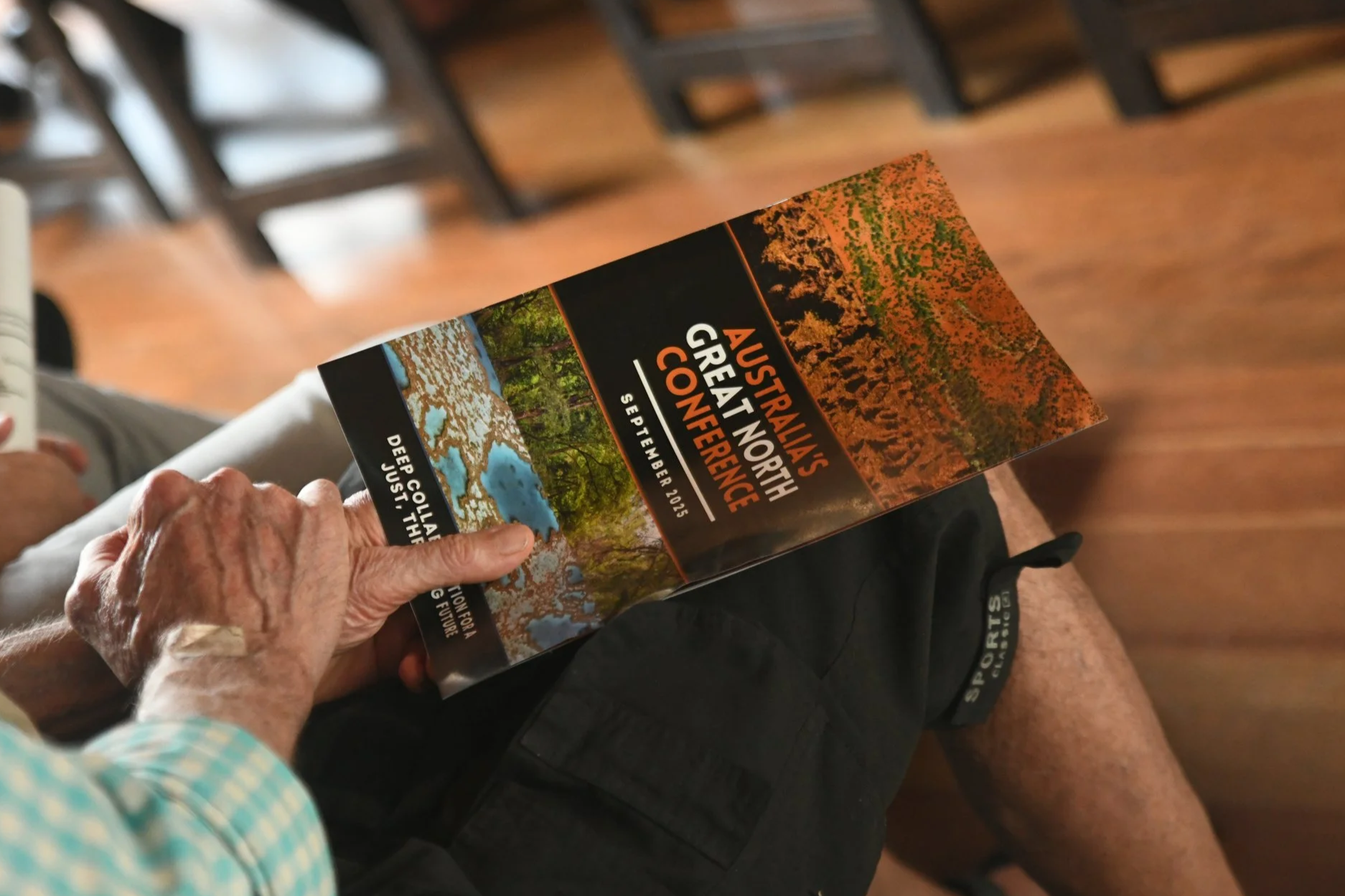 Close-up of an elderly person's hand holding a brochure titled 'Australia's Great North Conference,' with images of forests, lakes, and trees, and a date of September 2025.