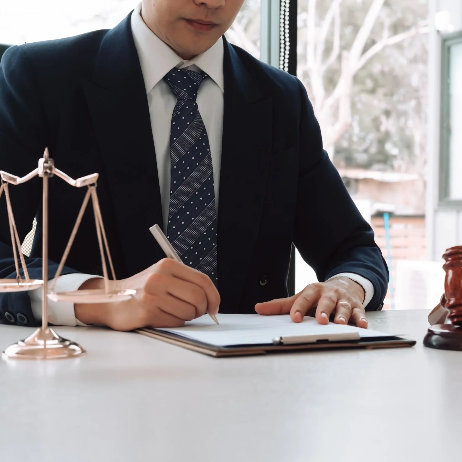 Person in a dark suit and striped tie writing on a clipboard, with a small balance scale and a gavel on the table.