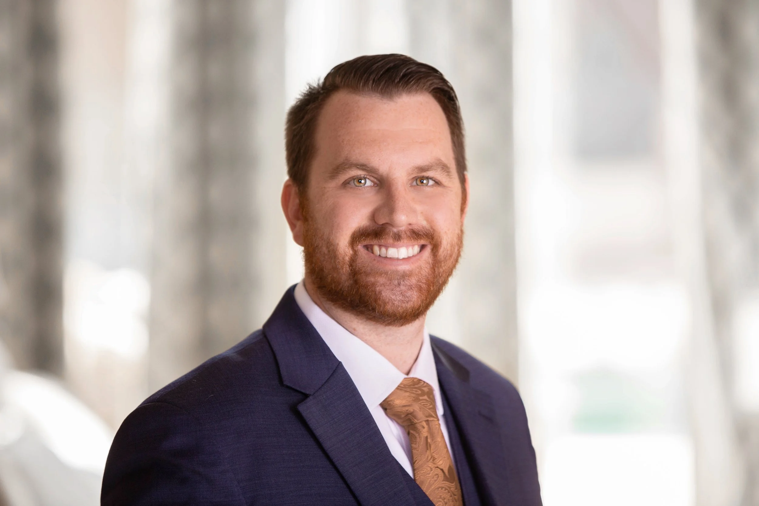 Portrait of a smiling man in a navy suit, white shirt, and gold tie, with a blurred background.