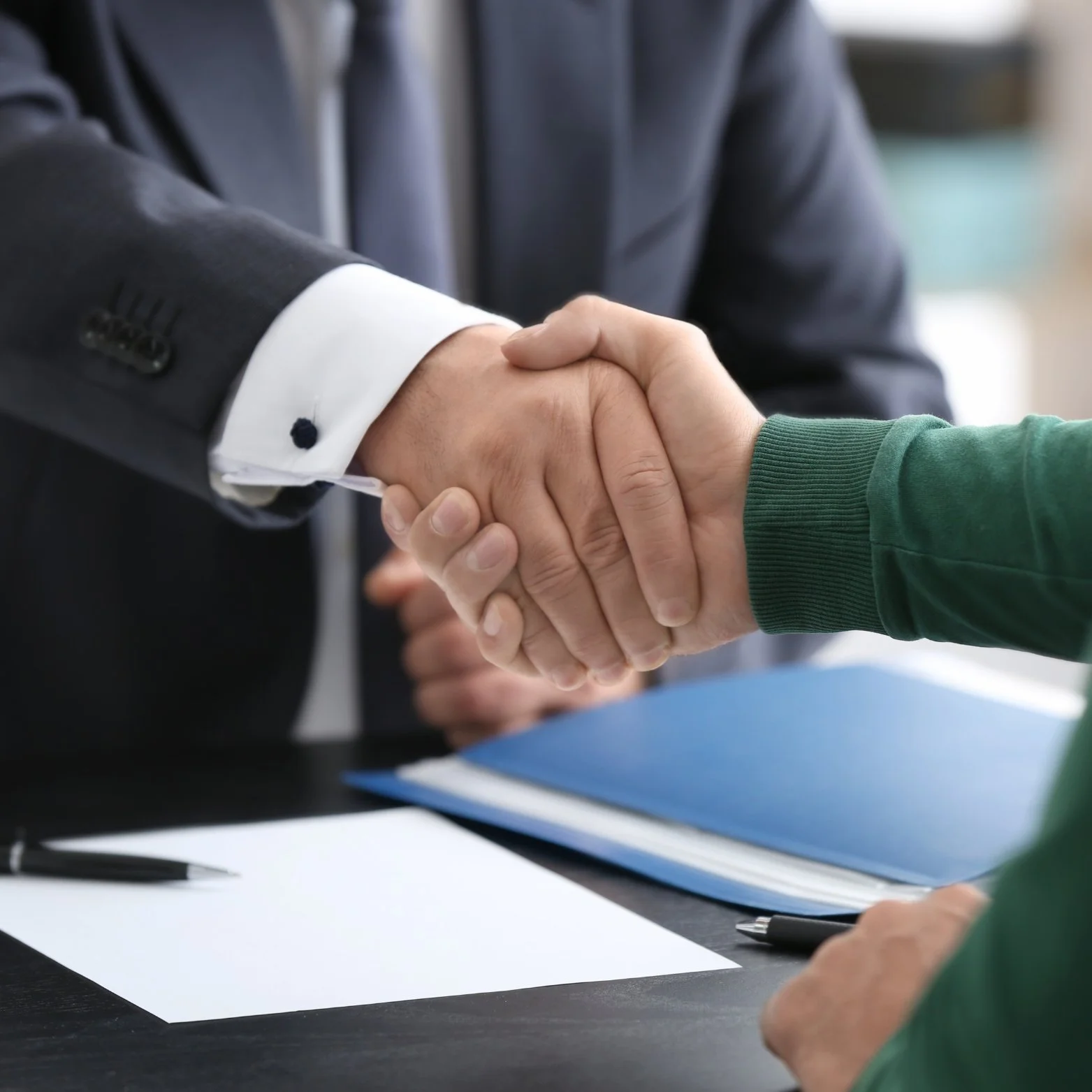 Close-up of two people shaking hands over a desk with documents and pens, indicating a business agreement or meeting.