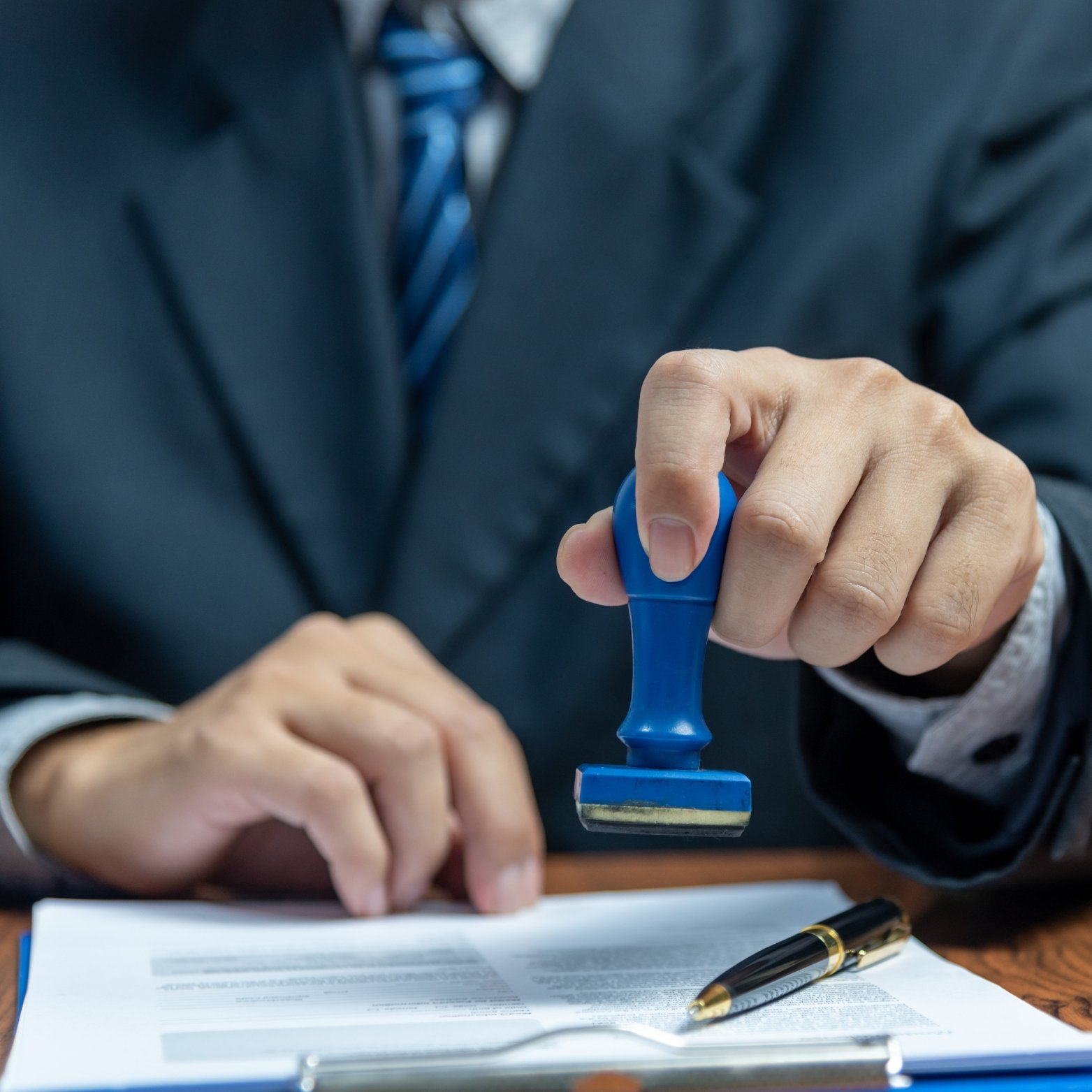 A person in a business suit stamping or pressing a document with a blue stamp, with a black and gold pen resting on the paper.