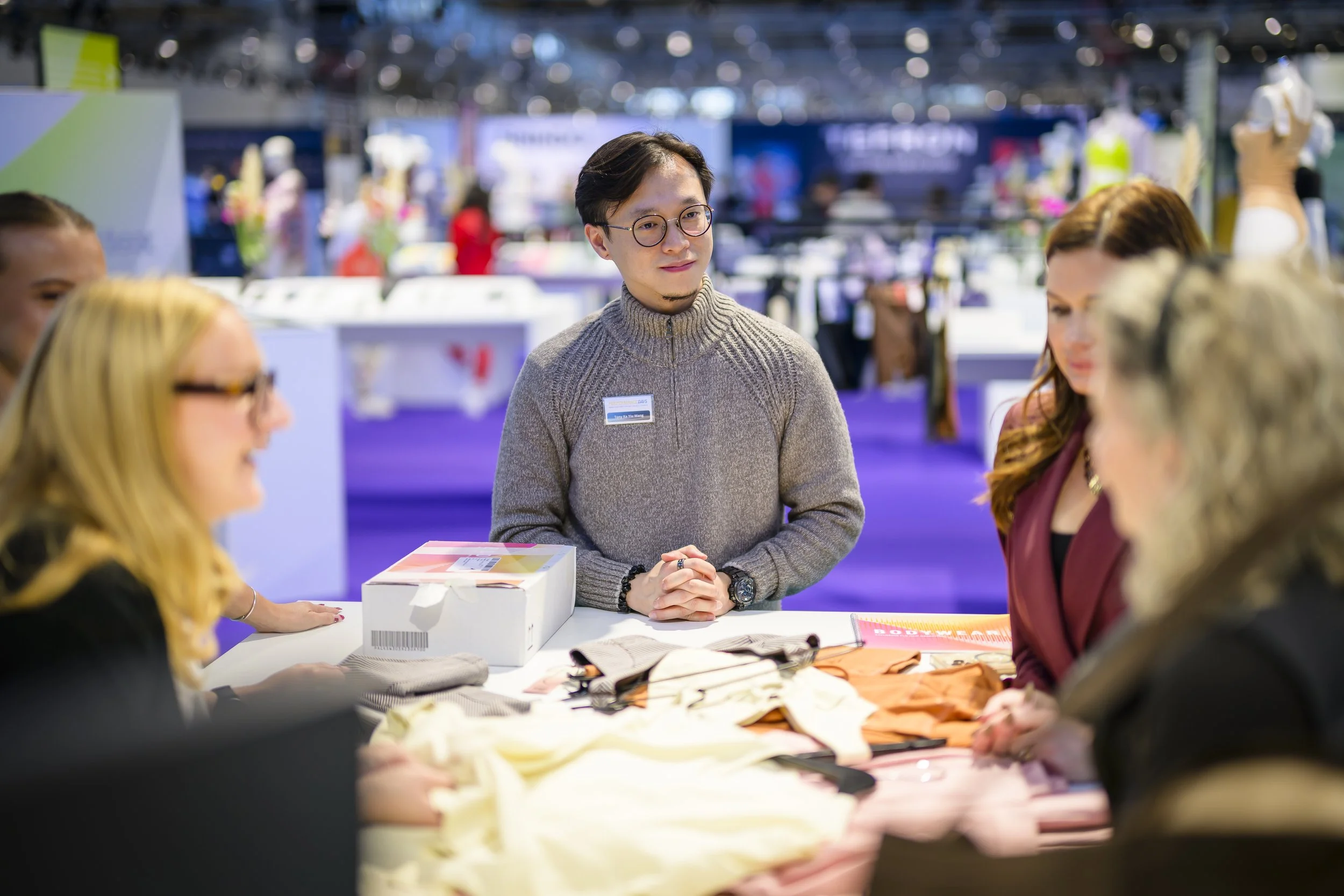 A man with glasses and a gray sweater standing at a table with people around him, in a brightly lit exhibition or trade show. The table has various items and a white box.