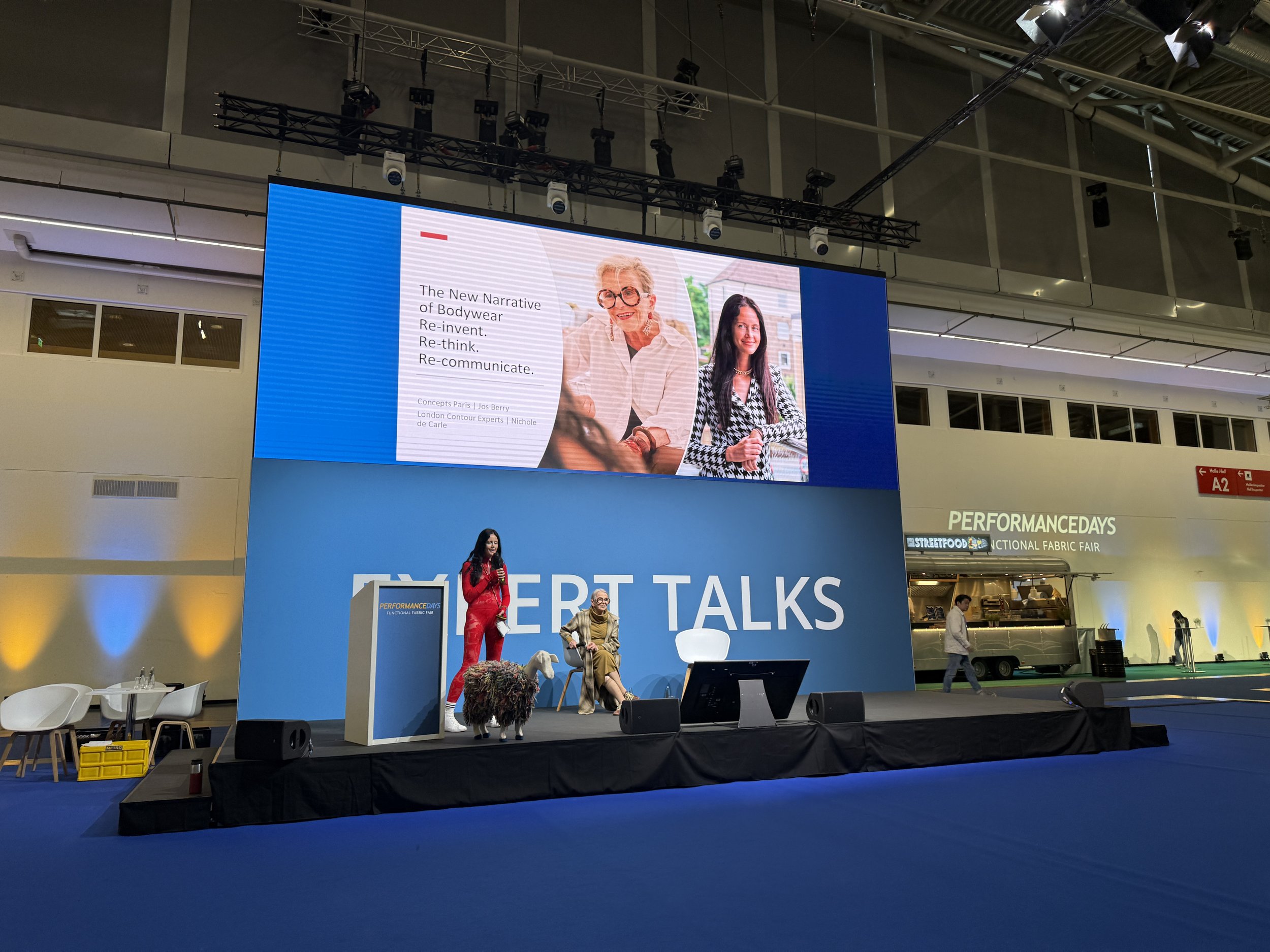 A presentation event with a stage featuring a large screen displaying a slide about bodywear, with two women pictured. In front, a woman in red is speaking at a podium, and another woman is seated on the stage with a woolly sheep costume.