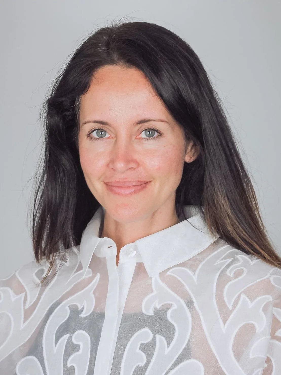 A woman with long dark hair and blue eyes wearing a white sheer blouse with floral embroidery, smiling at the camera.