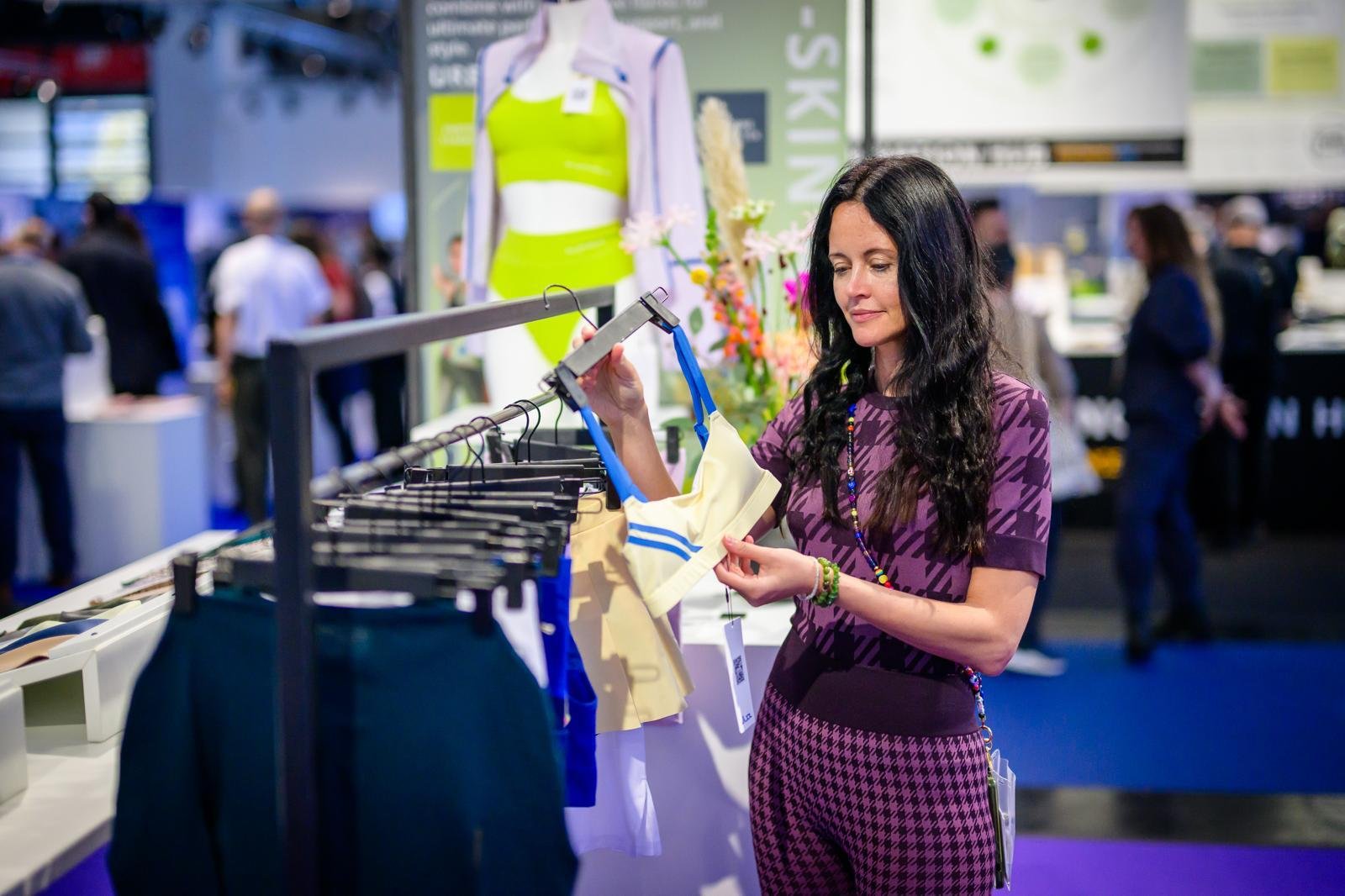 Woman shopping at a trade show or exhibition, browsing clothing items on a rack with other visitors in the background.