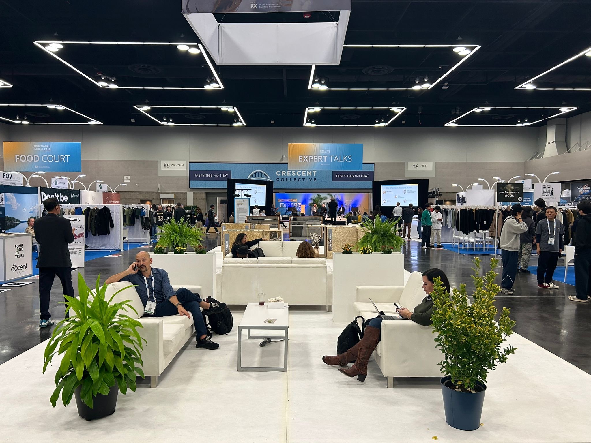 Event space with a central lounge area featuring white sofas, potted plants, and attendees seated using phones and laptops. In the background, there are booths and signs for food, expert talks, and various exhibitors at a trade show or conference.