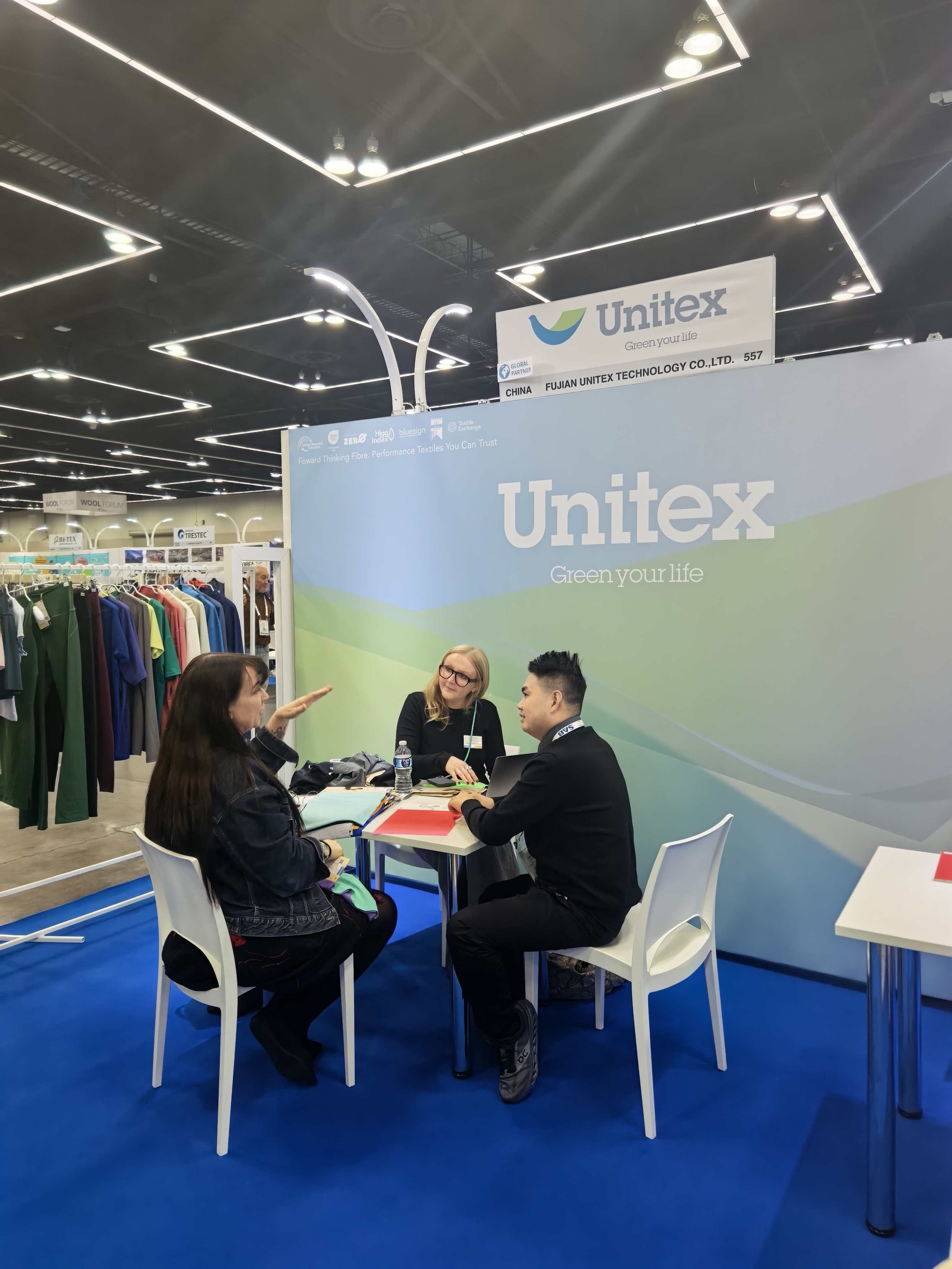 Three people seated around a table at an exhibition booth for Unitek, a textile company. They are engaged in a discussion with fabric samples and a laptop on the table. The booth has a large backdrop with the Unitek logo and slogan 'Green your life'.