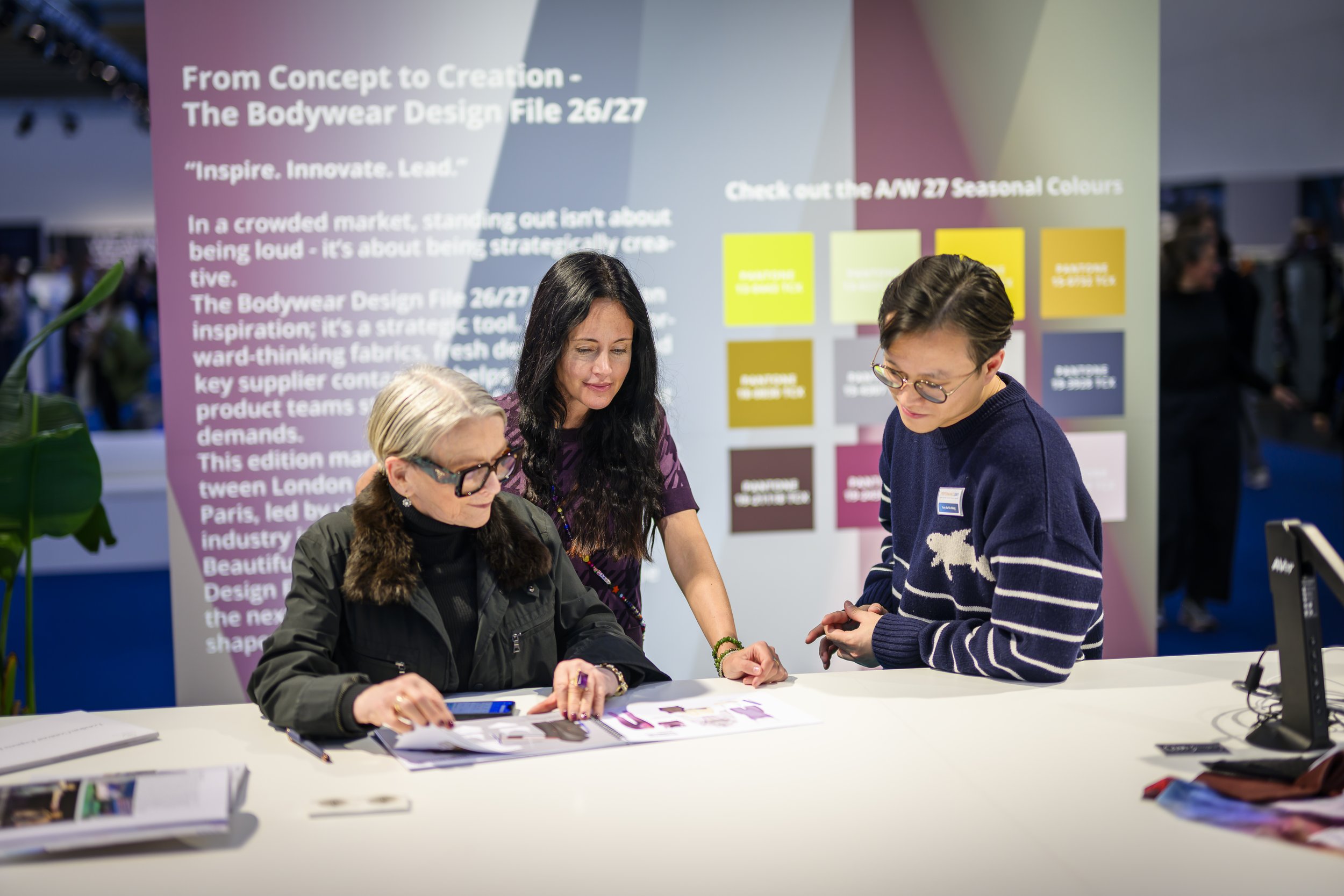 Three women are discussing design samples at a booth with a background poster about bodywear design and color options.