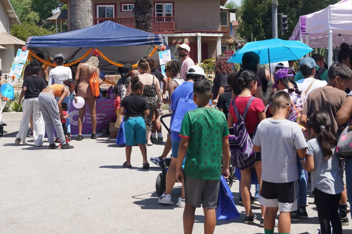 People gathered at a San Bernardino County Black Pride™ event with booths and decorations, some holding umbrellas for shade, on a sunny day in a residential area.