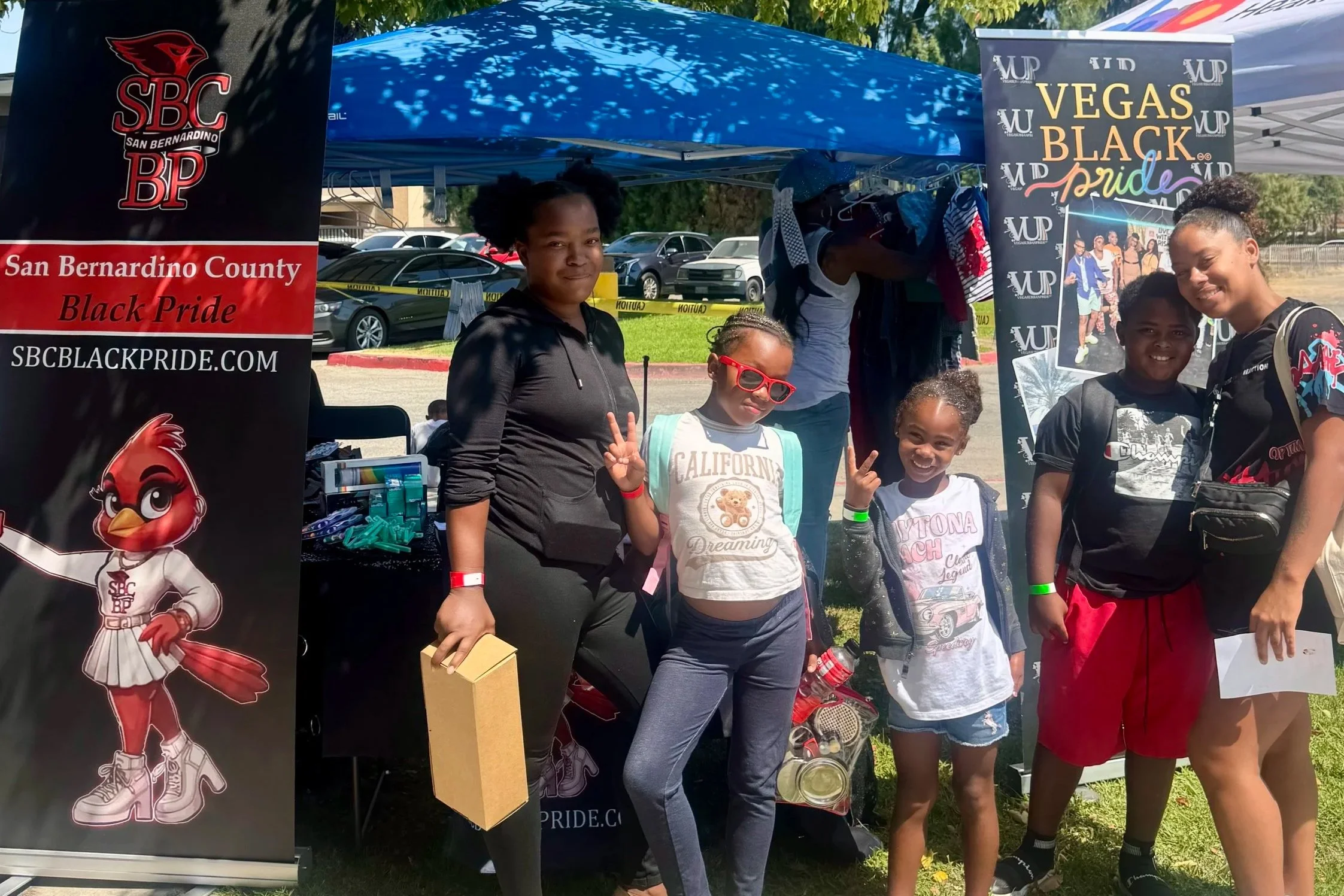 A group of people, including children and a woman, standing outdoors at a community event with banners promoting Black Pride in San Bernardino County Black Pride™ and Vegas Black Pride®. The background shows parked cars, trees, and a blue canopy.