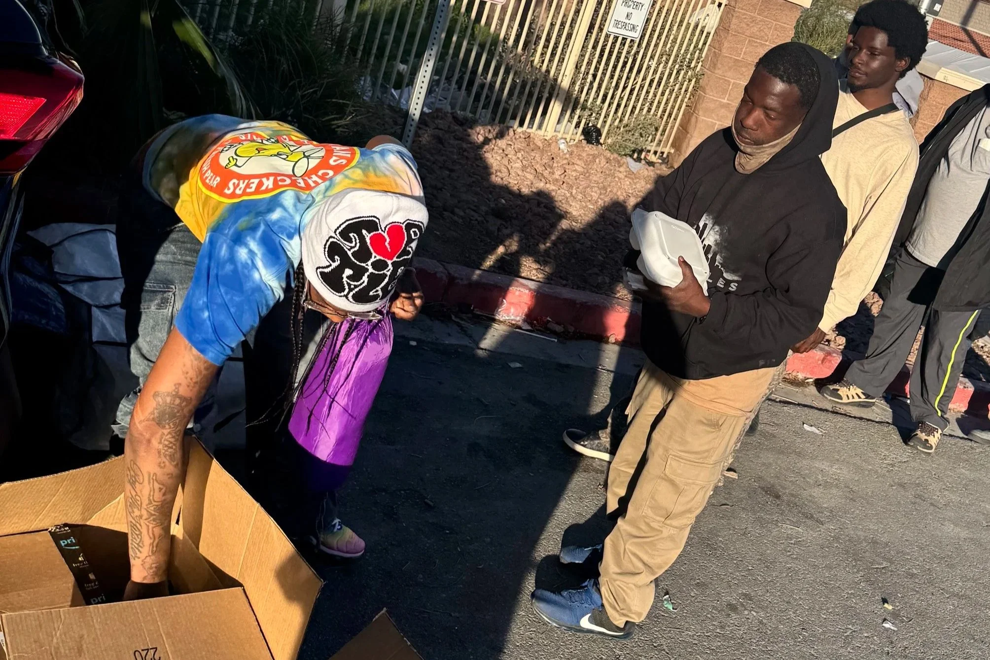 Group of people standing outdoors near a fence, with one person wearing a hoodie and another with a colorful tie-dye shirt and a hat, interacting around a cardboard box with items inside. Love Thy Neighbor program