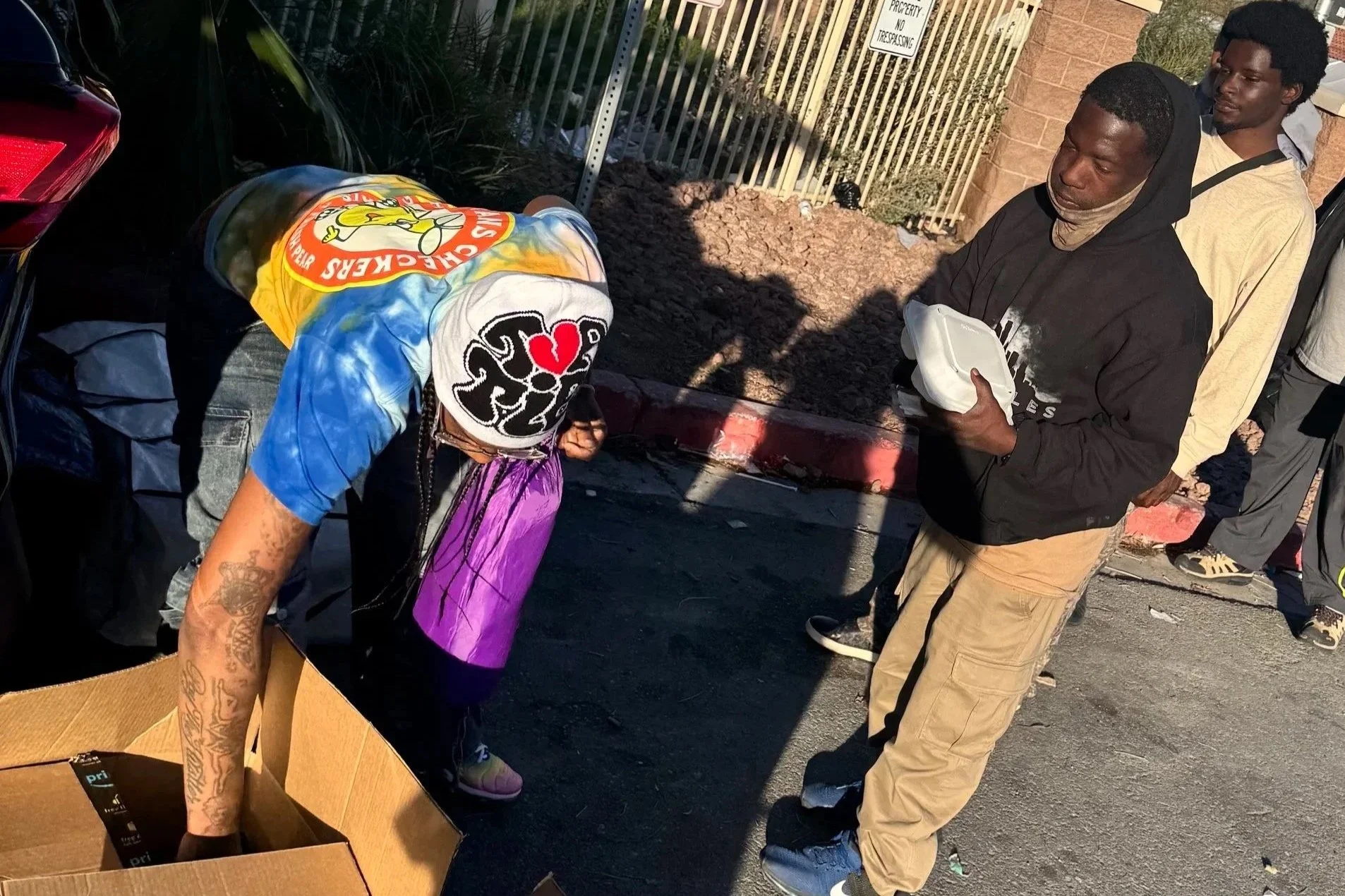 Love Thy Neighbor program Two young men, one wearing a black hoodie and tan pants, the other with a bandana on his face, are standing on a street near a parked car and a trash can. One is holding a foam takeout container.