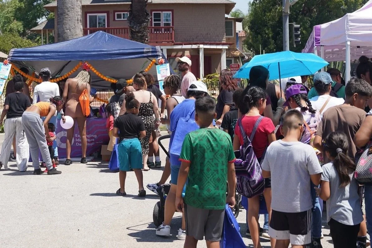 A crowd of people gathered under and around tents at an outdoor  San Bernardino County Black Pride™ event during the daytime. There are children and adults, some carrying bags or umbrellas, with a house or building in the background.
