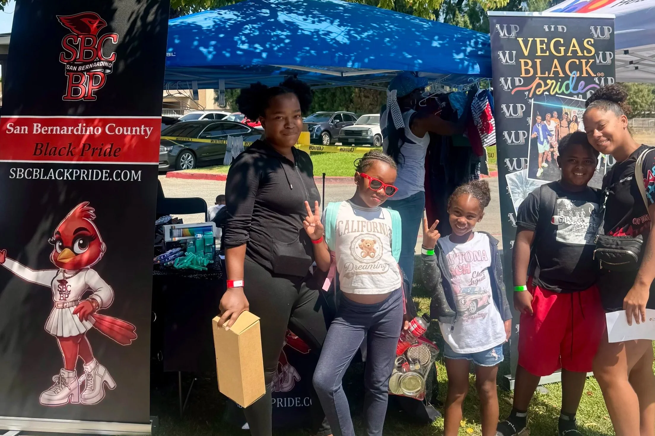 A group of people posing in front of promotional banners at an outdoor event. The banners promote San Bernardino County Black Pride Veags Black Pride. The group includes three women and three young girls, all smiling. The background shows parked cars and green trees.