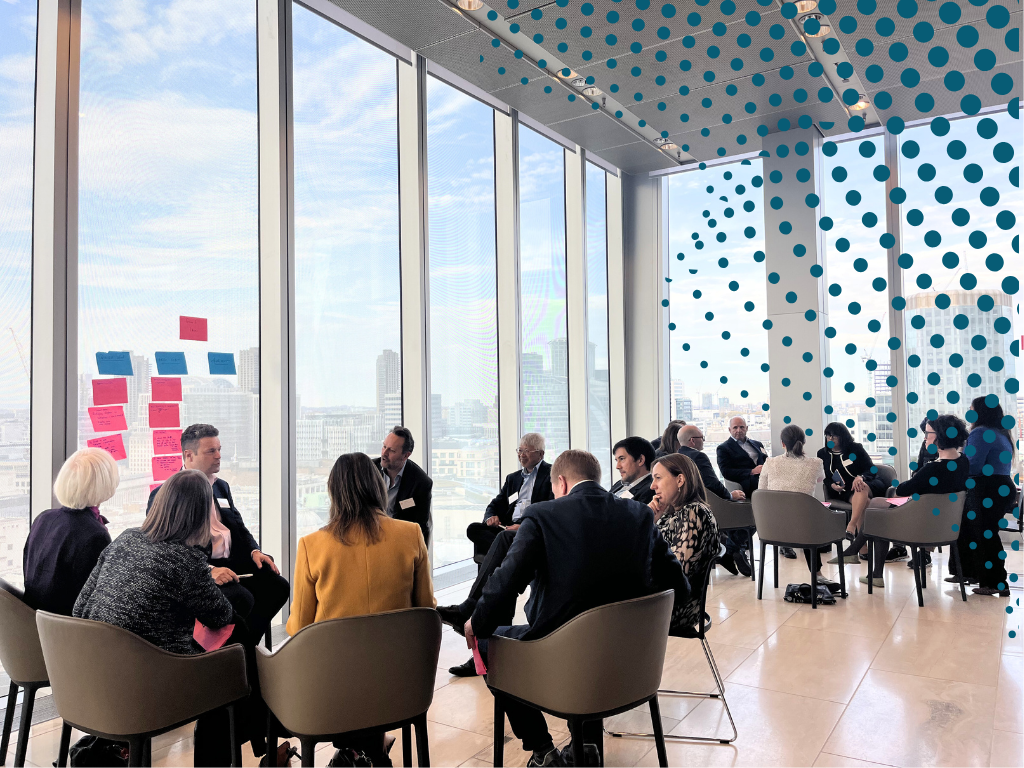 Image of people in small research groups around tables in front of a glass window with a city scape in the background.