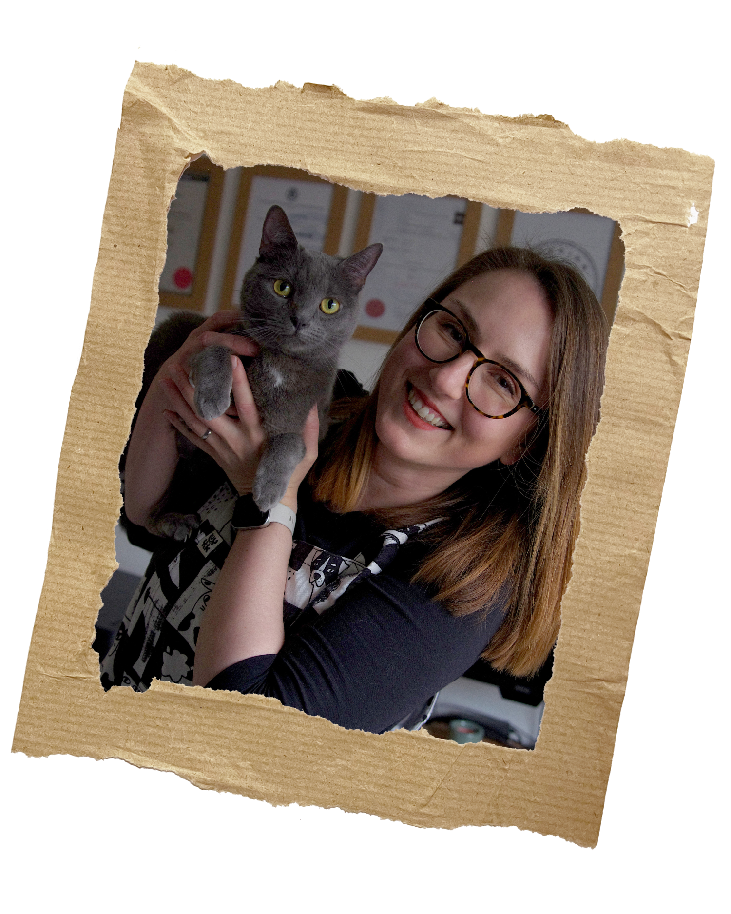 Anikó is holding her grey cat, Kiki up to the camera, she has a huge smile. In the background all her diplomas are displayed on the wall.