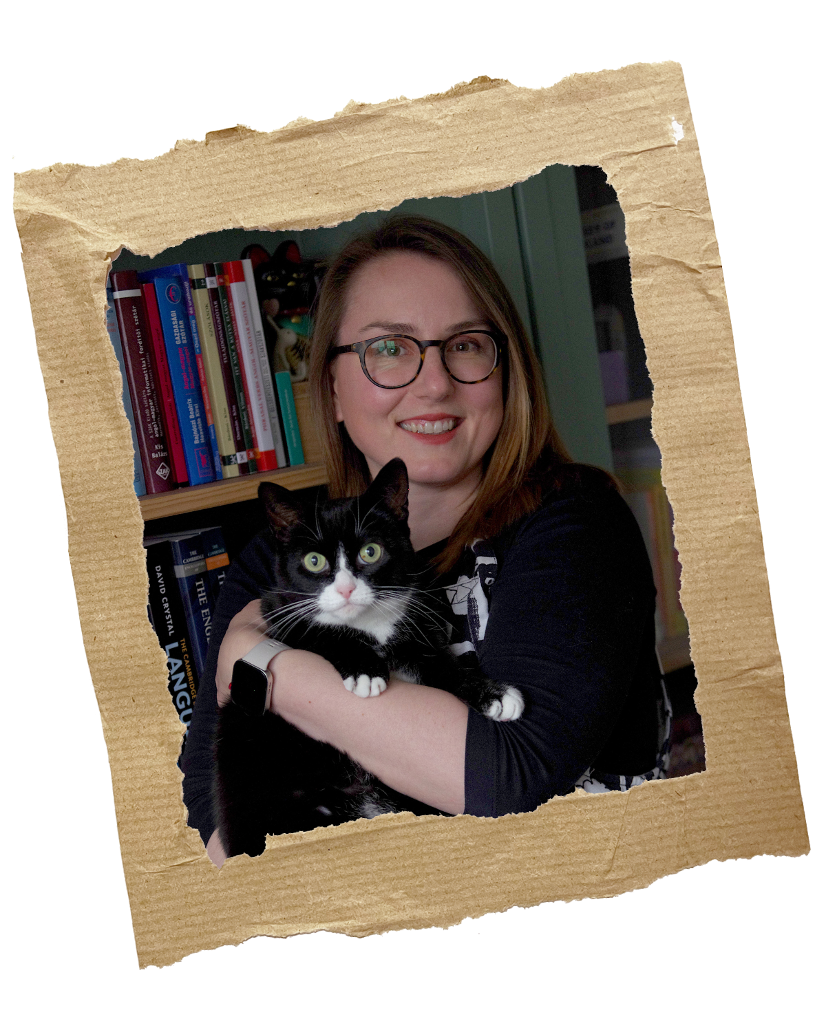 Anikó holding a tuxedo cat called Pip in her arms in front of a bookshelf. She's wearing a black top with black-white dungarees and a pair of glasses.