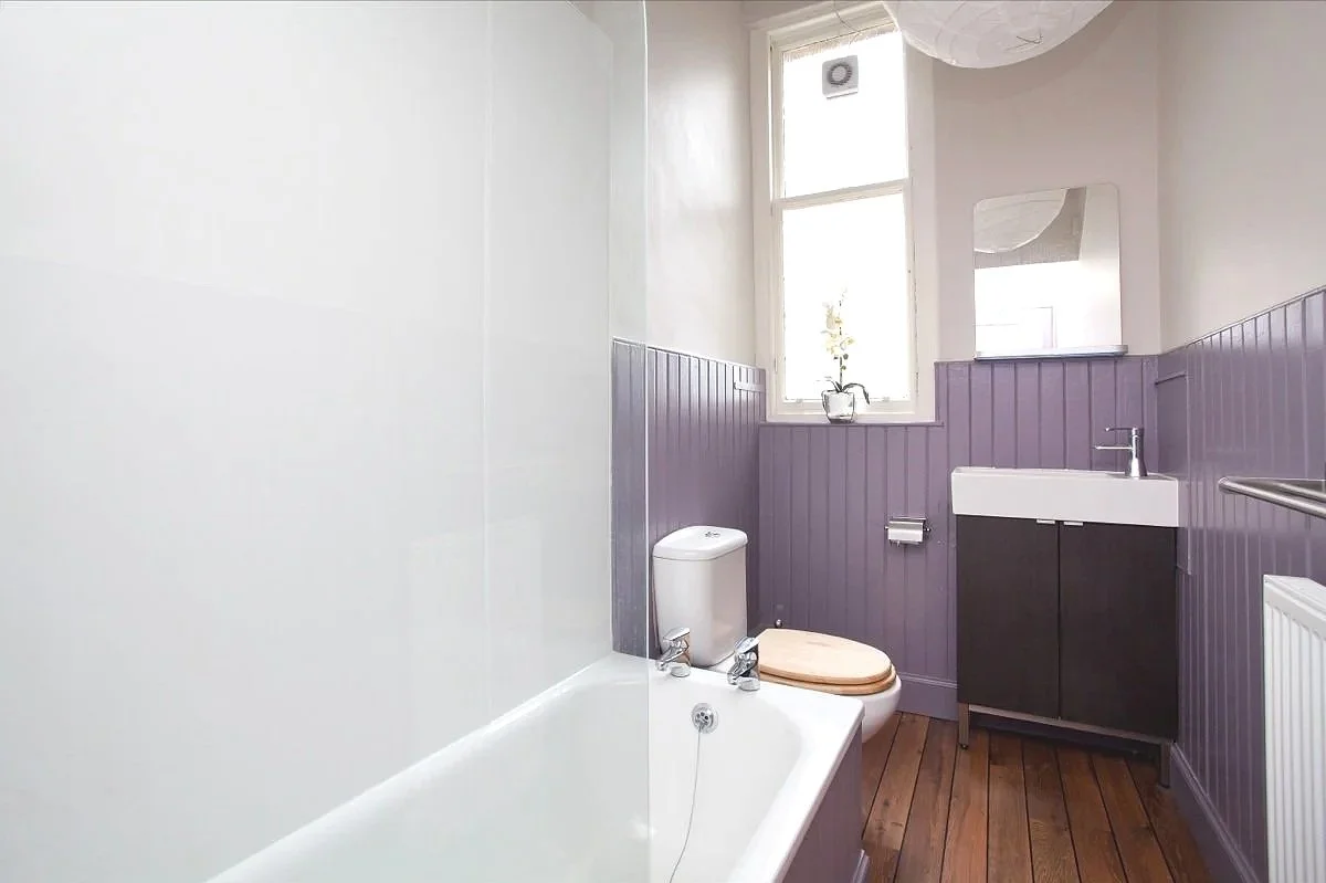 A small bathroom with purple beadboard wainscoting, a white bathtub with a wooden seat, a toilet, a small sink cabinet, a window with a plant, and a mirror on the wall.