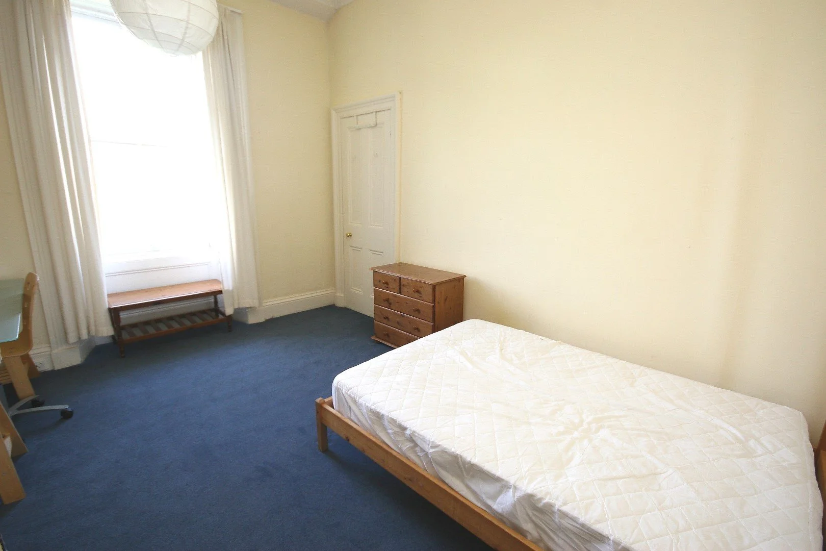 A sparsely furnished bedroom with a bare mattress on a wooden bed frame, a small wooden dresser, a window with white curtains, and a blue carpet.