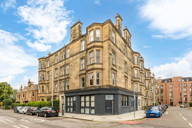 A corner view of a sandstone multi-story apartment building with bay windows, on a city street with parked cars and a blue sky with clouds.