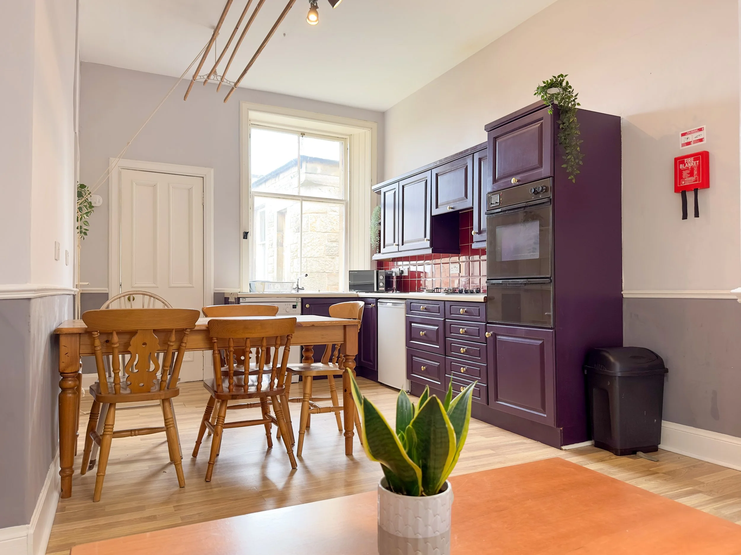 Bright kitchen with purple cabinets, wooden dining table with four chairs, potted plant on counter, window letting in natural light, red tiled backsplash, and a fire alarm on the wall.