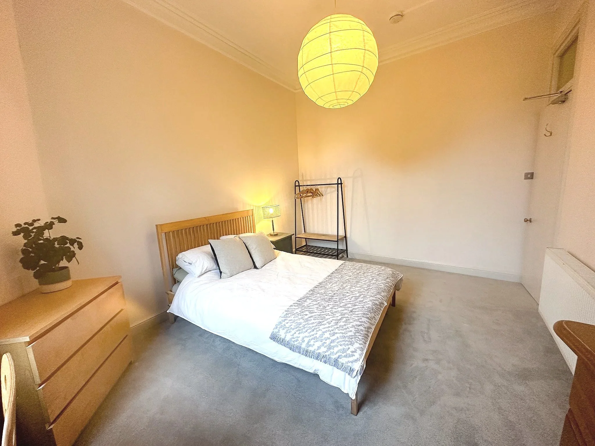 A bedroom with a wooden bed frame, white bedding, and gray pillows, a wooden dresser with a potted plant, a nightstand with a lamp, a clothing rack, and a large paper lantern ceiling light.