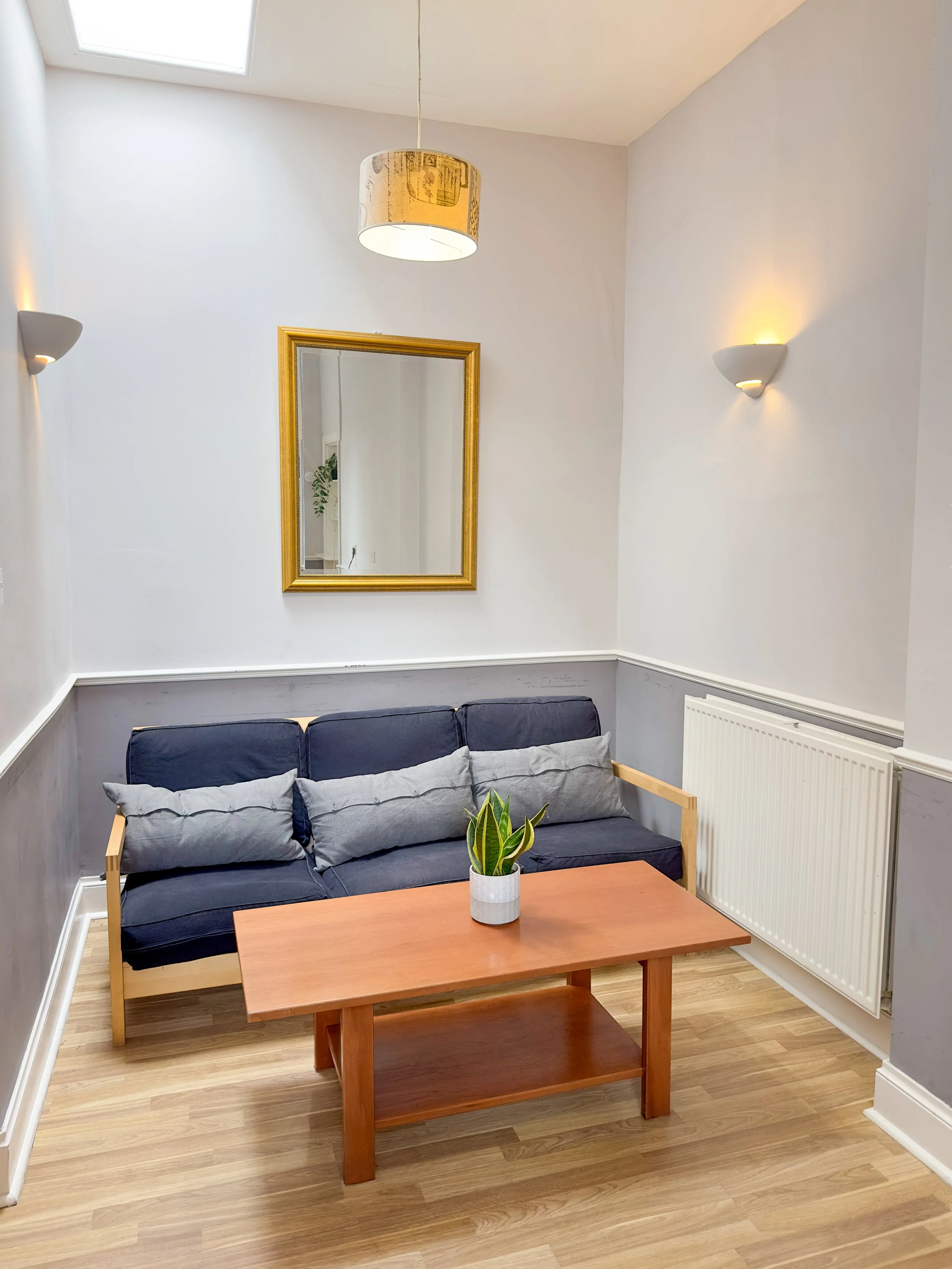 A small living area with a navy blue sofa, wooden coffee table, and a potted plant on the table. There is a mirror with a gold frame on the wall, two wall sconces, and a ceiling light fixture. The room has white walls and a hardwood floor.
