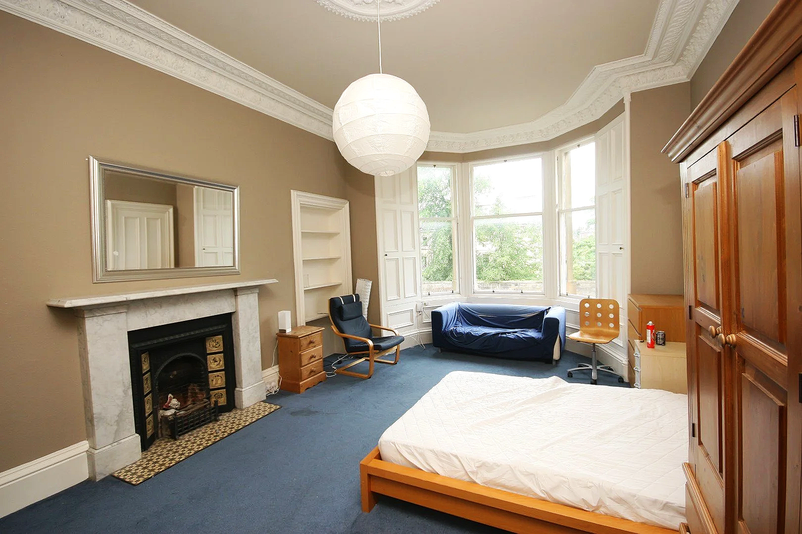 A bedroom with beige walls, a white fireplace, a large bay window, and a blue carpet. There is a bed with a white mattress, a wooden wardrobe, a wooden chair, a sofa covered with a blue sheet, a small wooden nightstand, and a ceiling light.